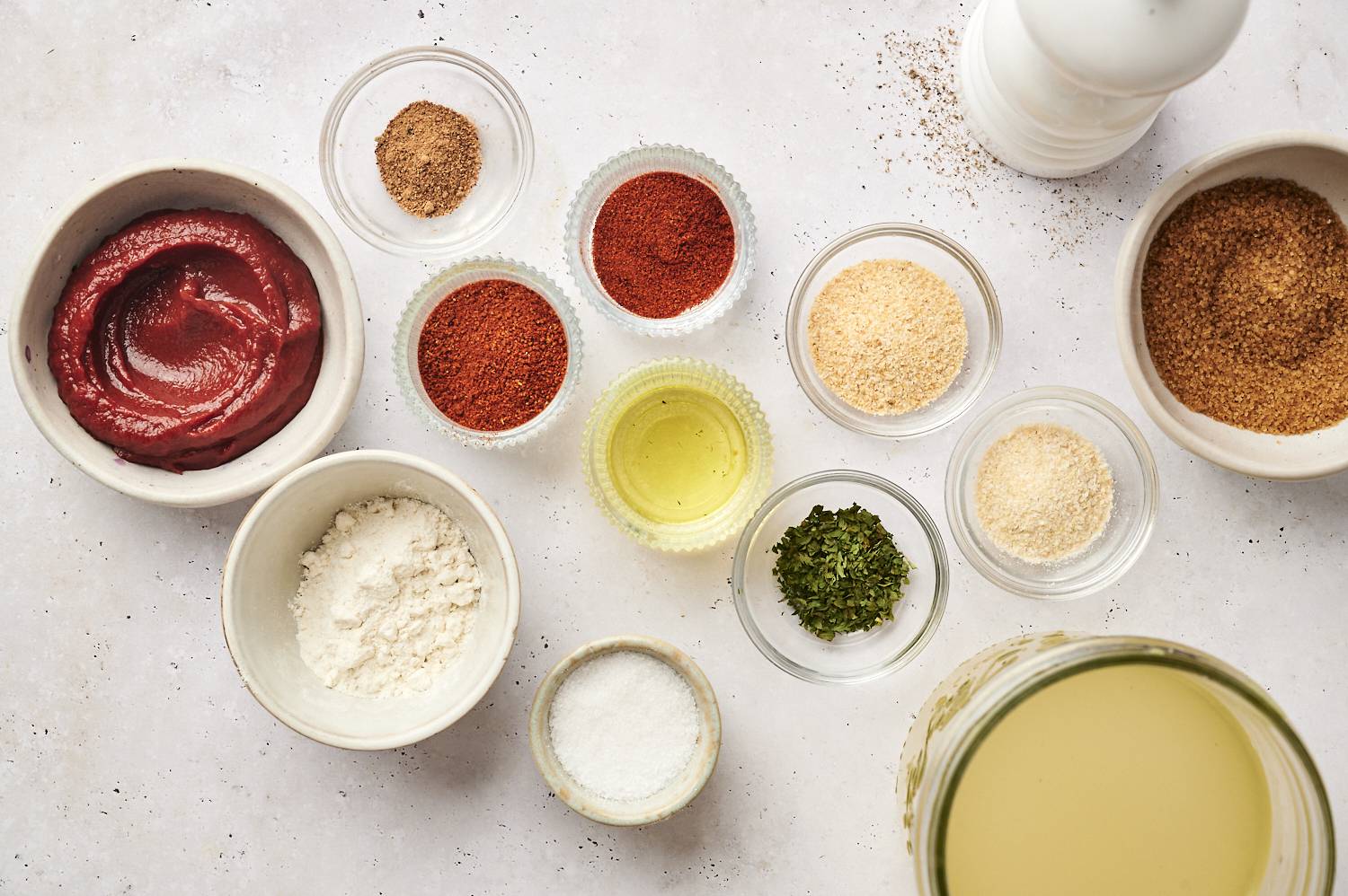 A flat-lay view of ingredients for homemade enchilada sauce, including tomato paste, flour, chili powder, garlic powder, onion powder, cumin, brown sugar, oil, broth, salt, and dried herbs, arranged in small bowls.