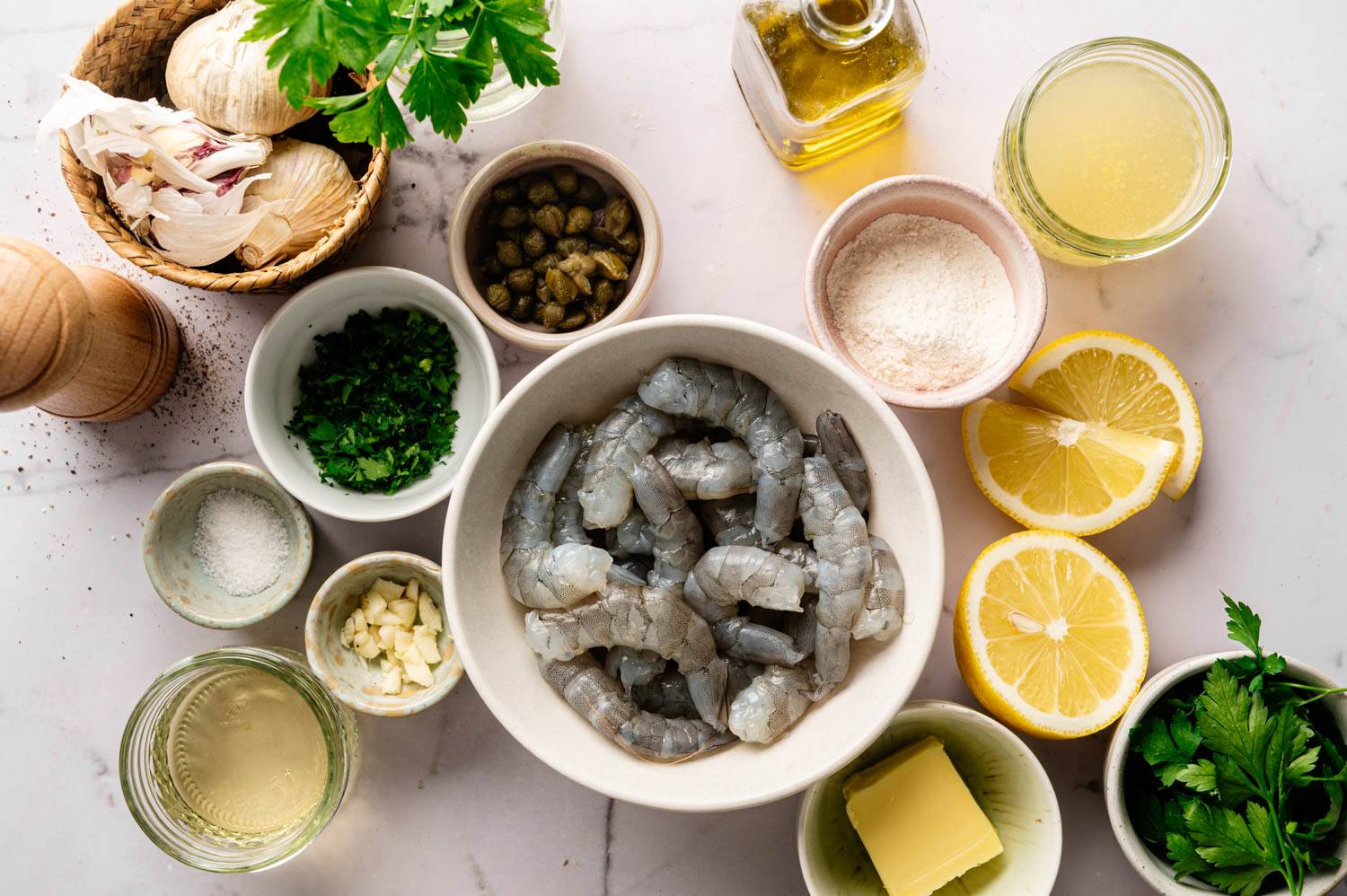 Top-view of raw shrimp in a bowl surrounded by ingredients: lemon slices, garlic, herbs, capers, olive oil, butter, flour, and a pepper mill on a white surface.
