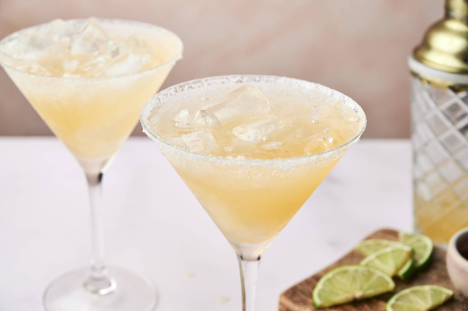 Two chilled cocktails in rimmed glasses with ice, beside lime wedges on a wooden board, set against a soft pink background.