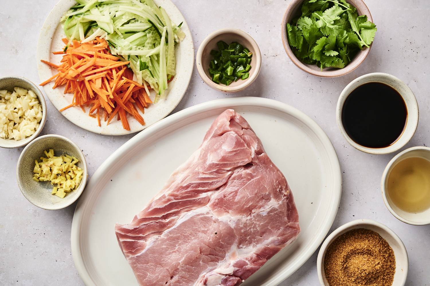 A raw pork slab surrounded by fresh veggies and seasonings, including garlic, ginger, cilantro, and sauces in small bowls.
