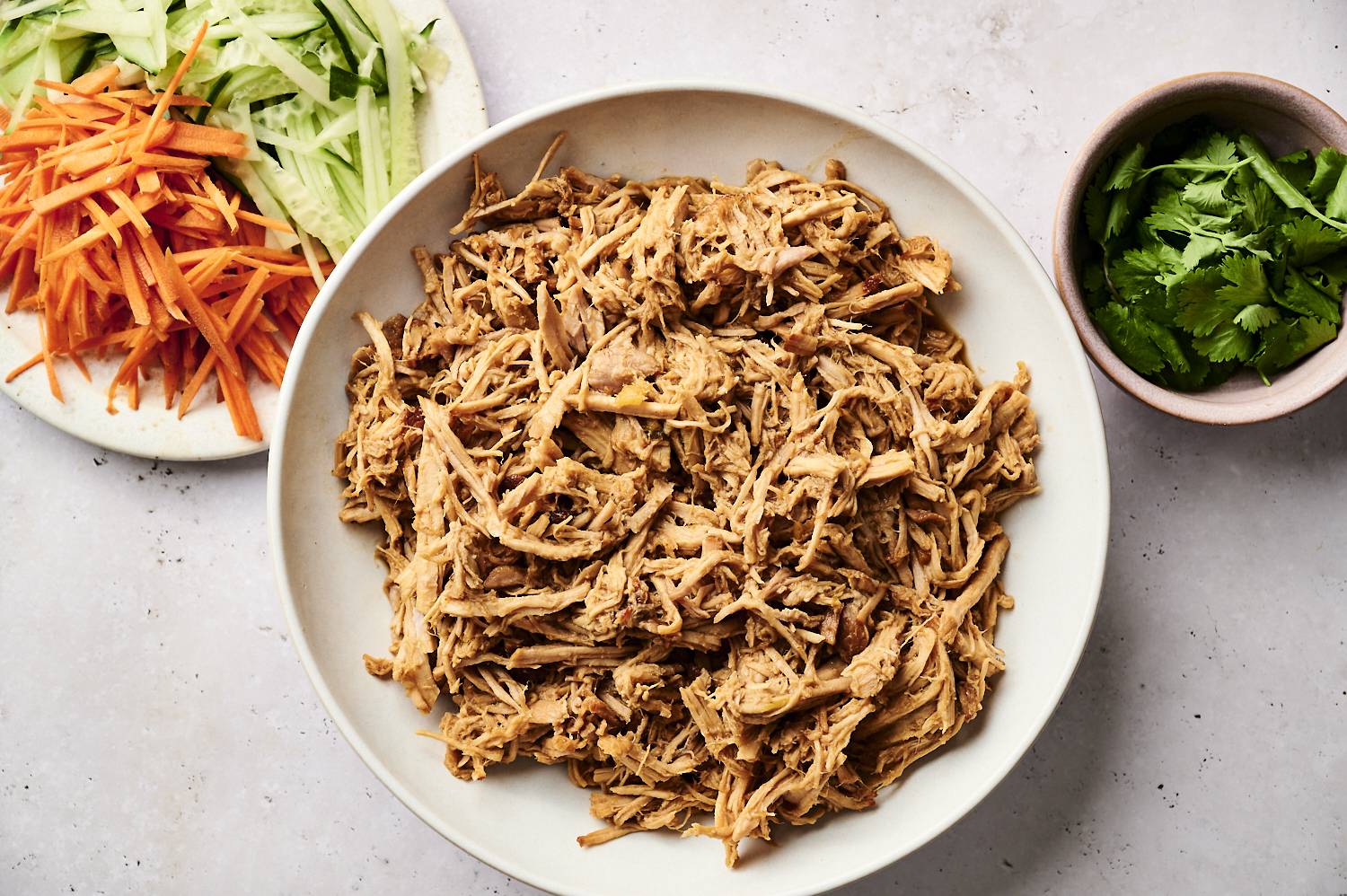 A bowl of shredded meat surrounded by sliced cucumbers, carrots, and fresh cilantro on a light marble surface.