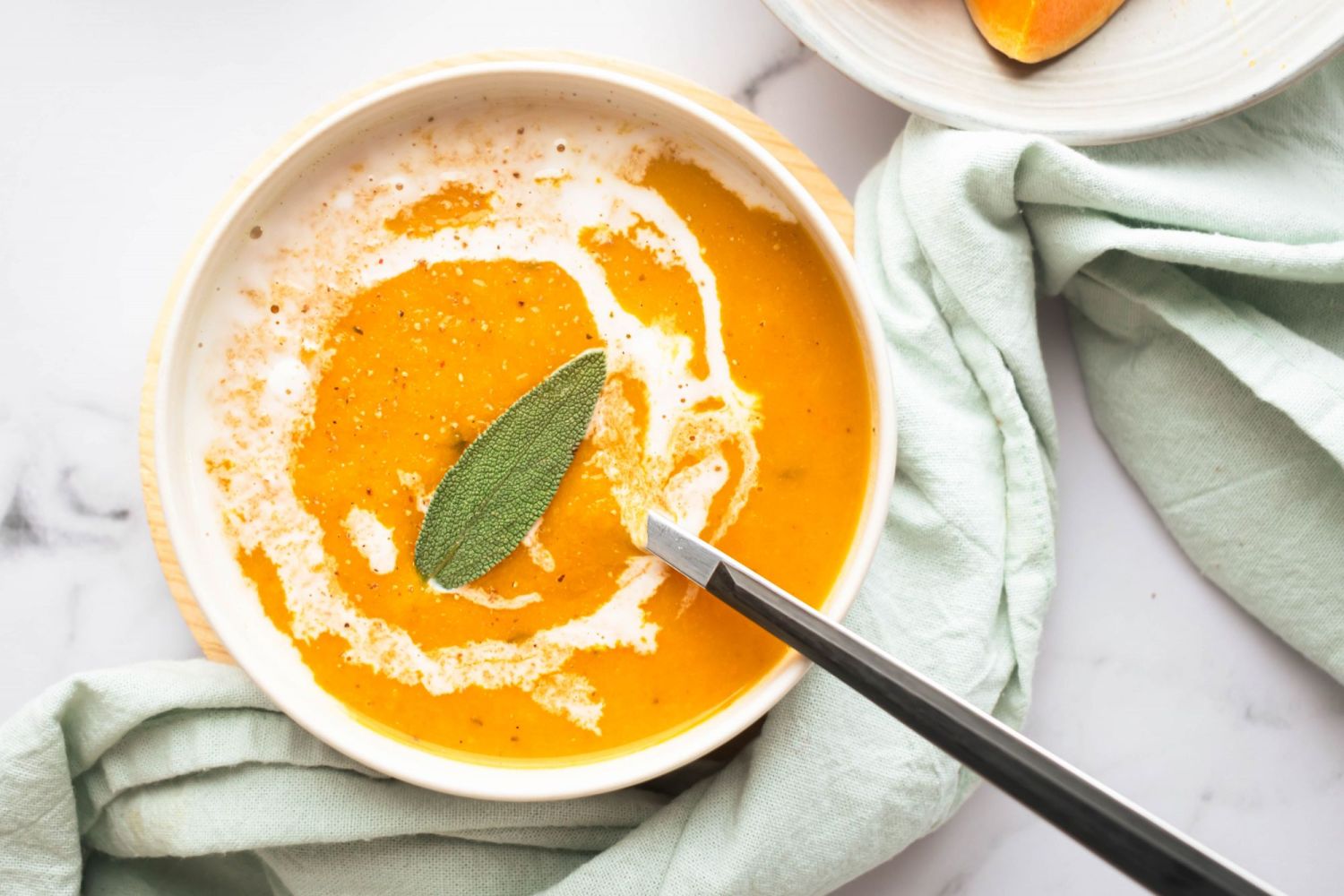 Butternut squash and apple soup in a bowl with a swirl of coconut milk, sage leaf, and light blue napkin.