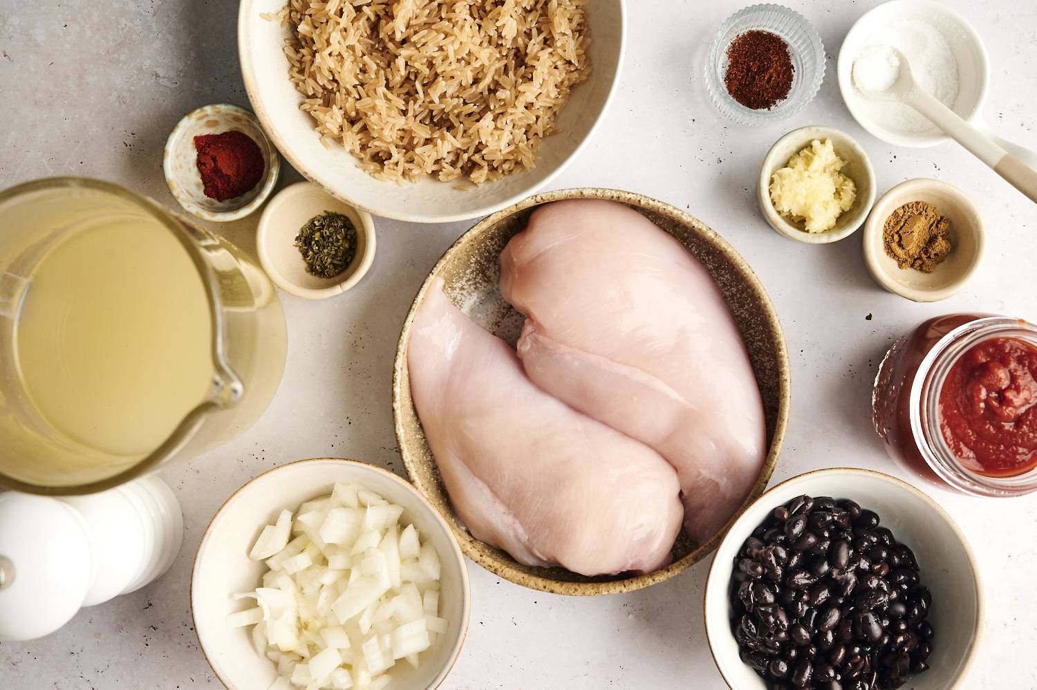A flat-lay view of ingredients for slow cooker chicken burrito bowls, including raw chicken breasts, black beans, rice, diced onions, chicken broth, tomato sauce, and various spices and seasonings arranged in bowls and jars.
