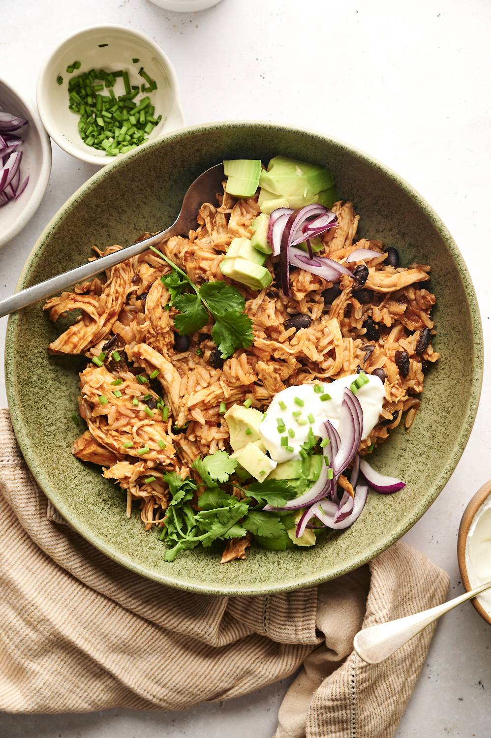 A top-down view of a bowl of chicken burrito rice topped with avocado, red onions, cilantro, sour cream, and chopped chives, served in a green ceramic dish with a spoon. Small bowls of extra toppings are arranged around the dish.