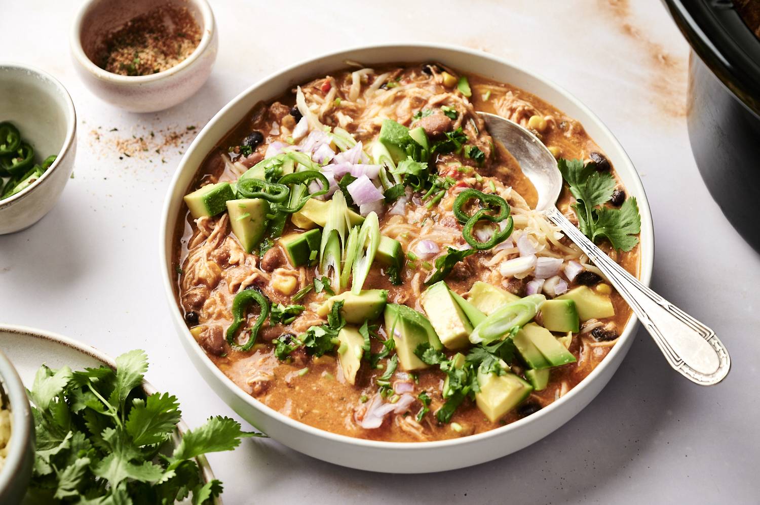 Bowl of chicken enchilada soup topped with diced avocado, jalapeño slices, red onions, green onions, cilantro, and shredded cheese, served with a spoon.