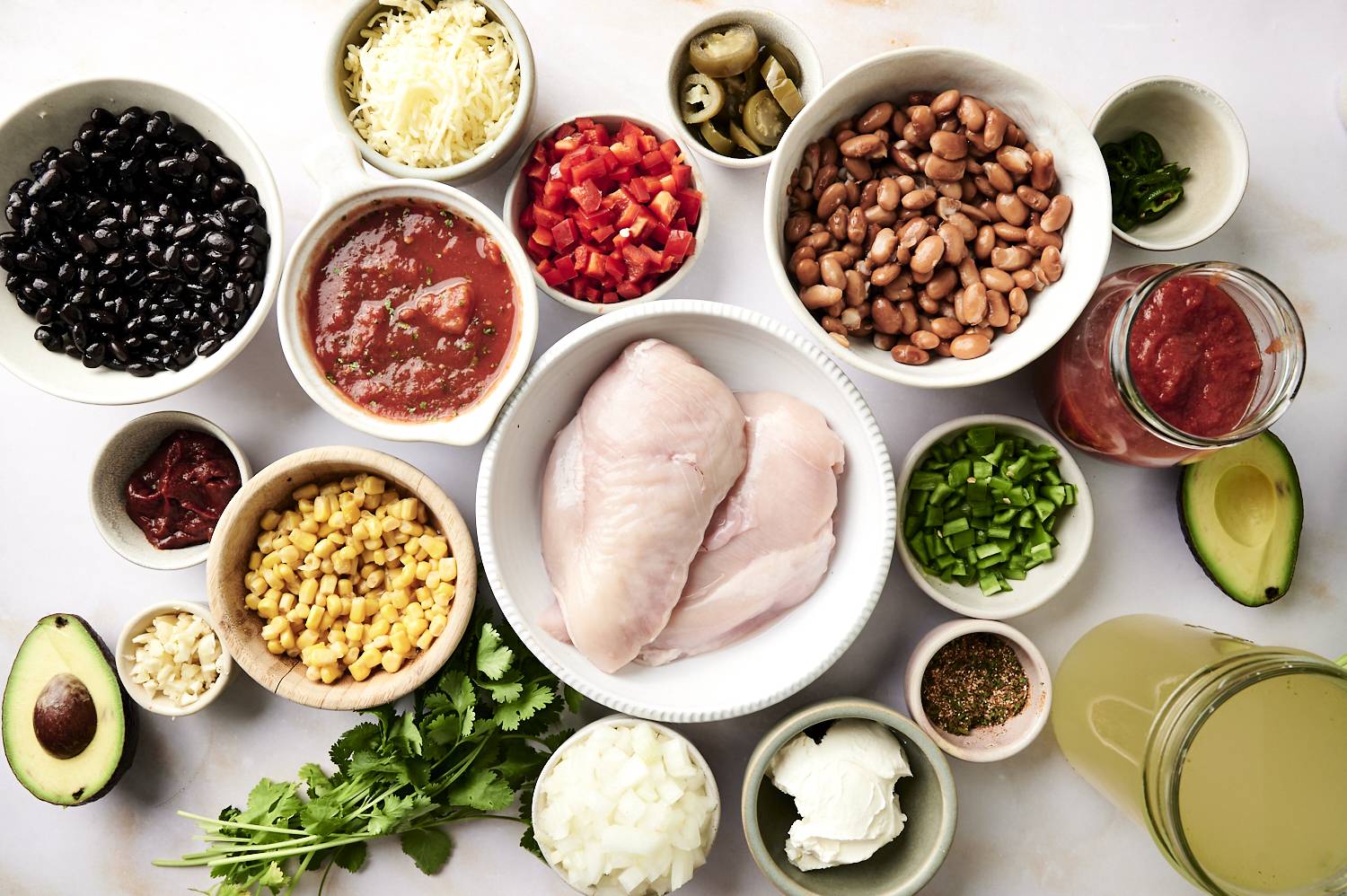 Ingredients for chicken enchilada soup including raw chicken breasts, black beans, pinto beans, corn, diced tomatoes, salsa, green chilies, jalapeños, cheese, avocado, chicken broth, and spices arranged in bowls.