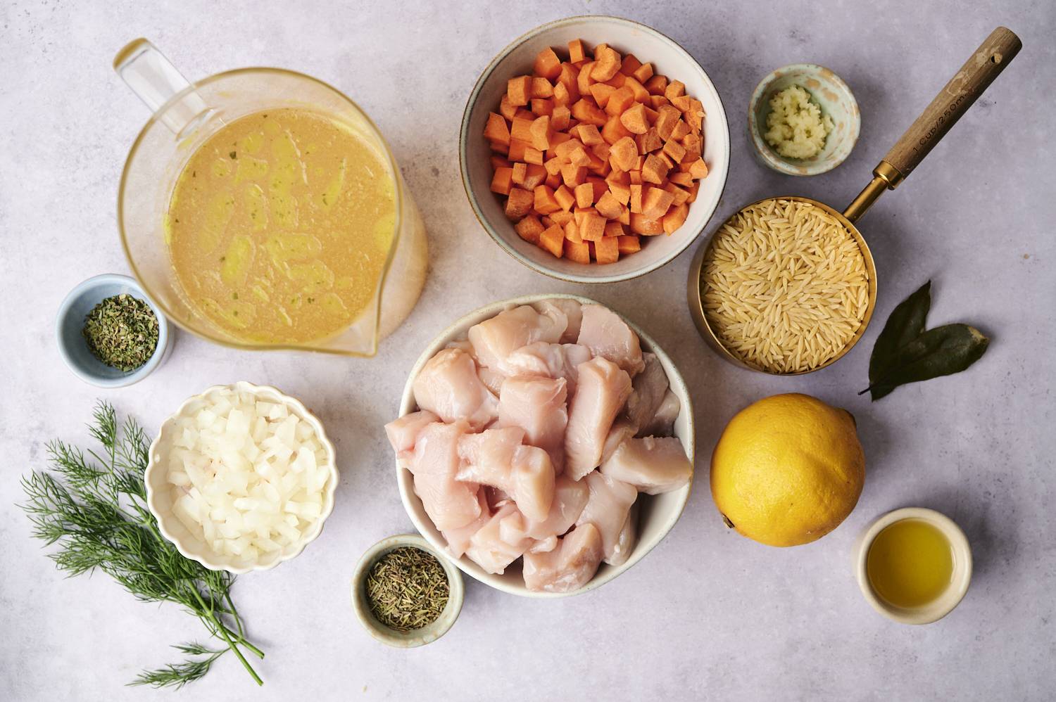 Ingredients including diced chicken, carrots, onions, orzo, broth, lemon, herbs, and olive oil arranged on a countertop.