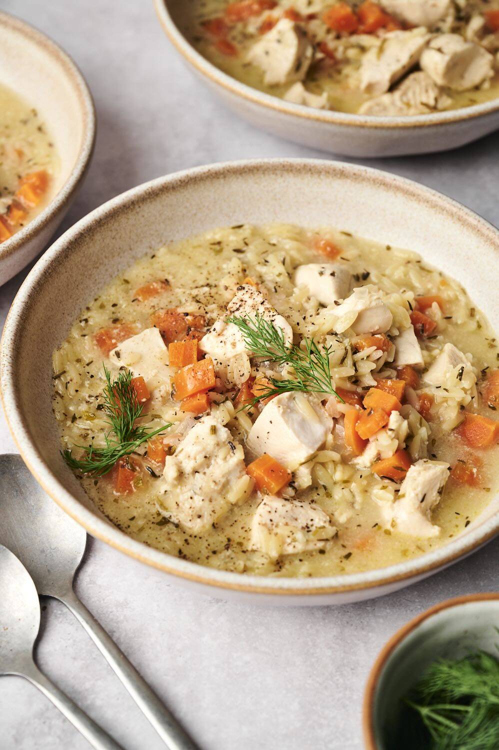A bowl of hearty chicken soup with orzo, carrots, and fresh dill, accompanied by two metal spoons and a bowl of fresh dill.