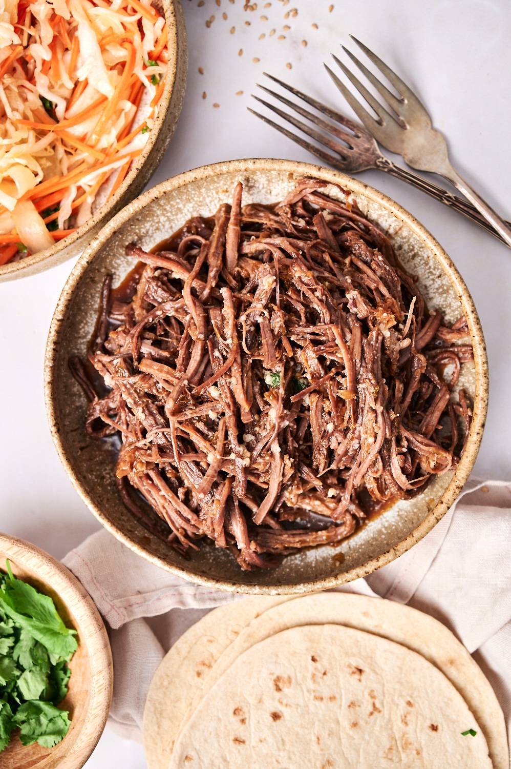Shredded Korean beef in a bowl for tacos with coleslaw, tortillas, and cilantro on the side.
