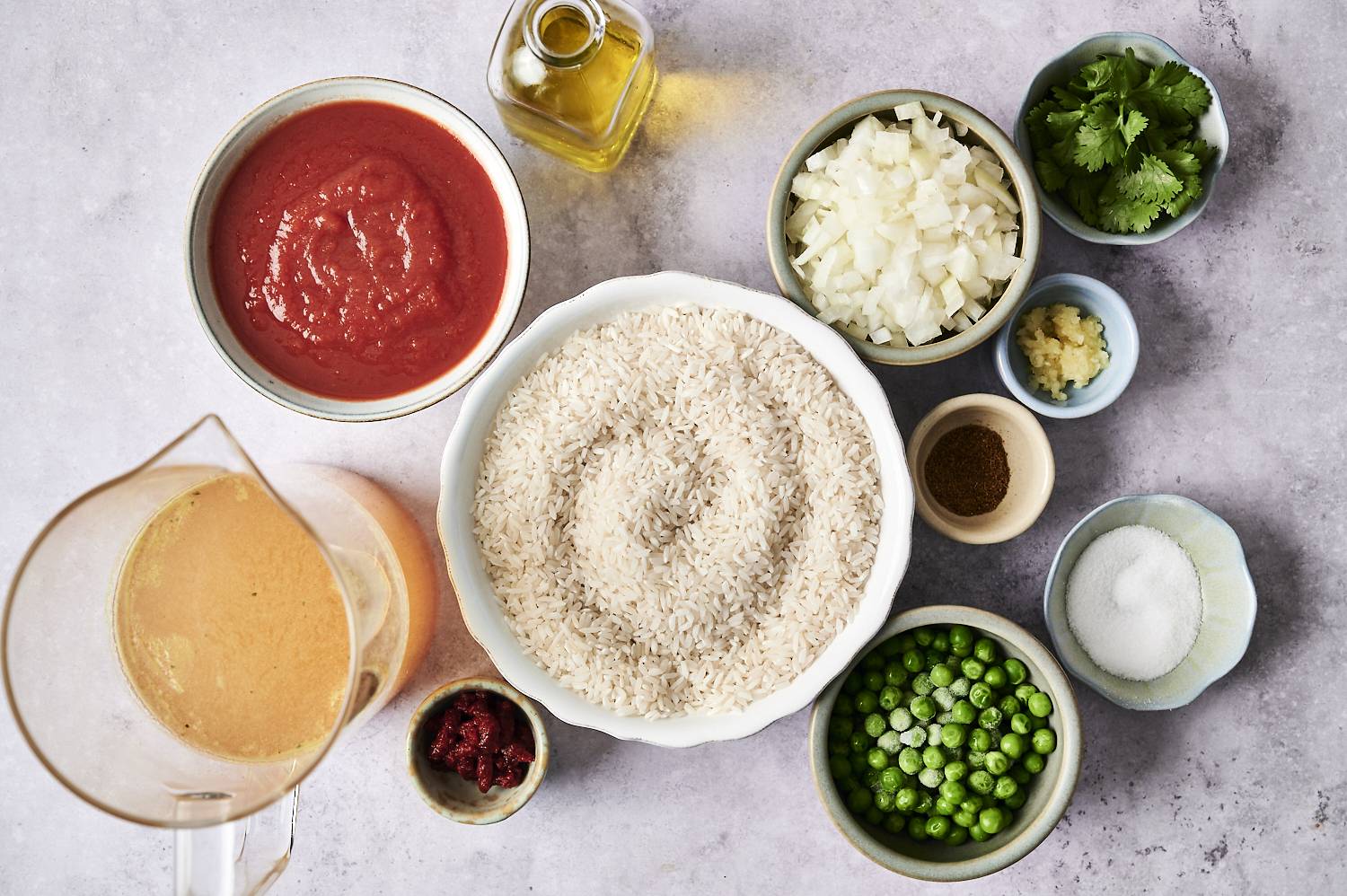Overhead view of ingredients for slow cooker Mexican rice, including a large bowl of uncooked white rice surrounded by bowls of tomato sauce, chopped onions, minced garlic, frozen peas, tomato paste, chicken broth, oil, chopped cilantro, and spices on a light gray surface.