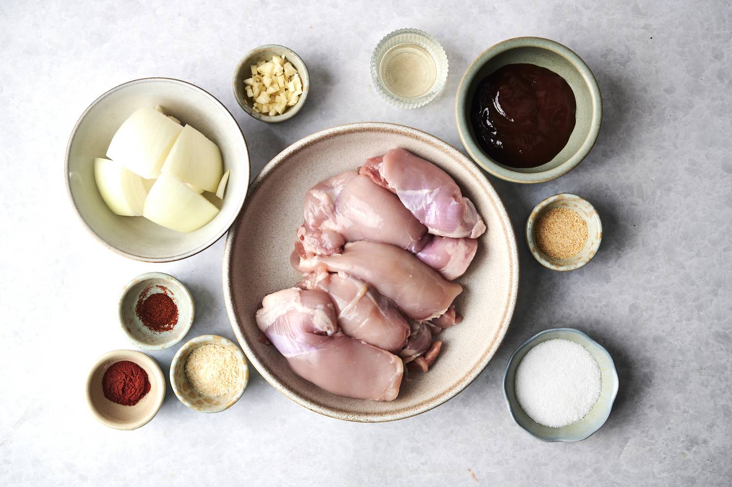 An array of cooking ingredients, including chicken, onions, spices, and sauces, arranged neatly on a gray surface.