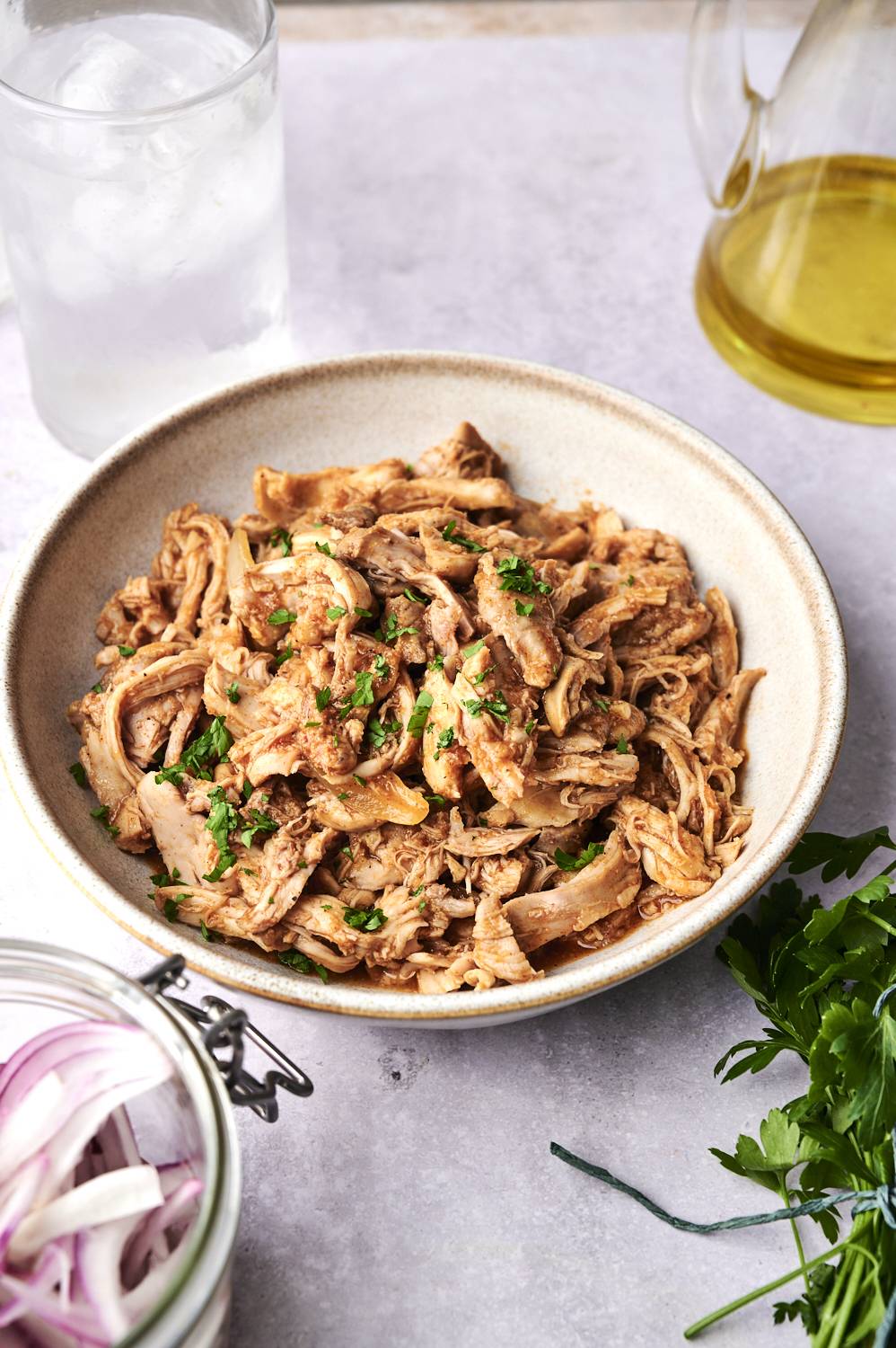 A bowl of shredded chicken garnished with parsley, accompanied by sliced onions, a glass of water, and olive oil on a gray background.