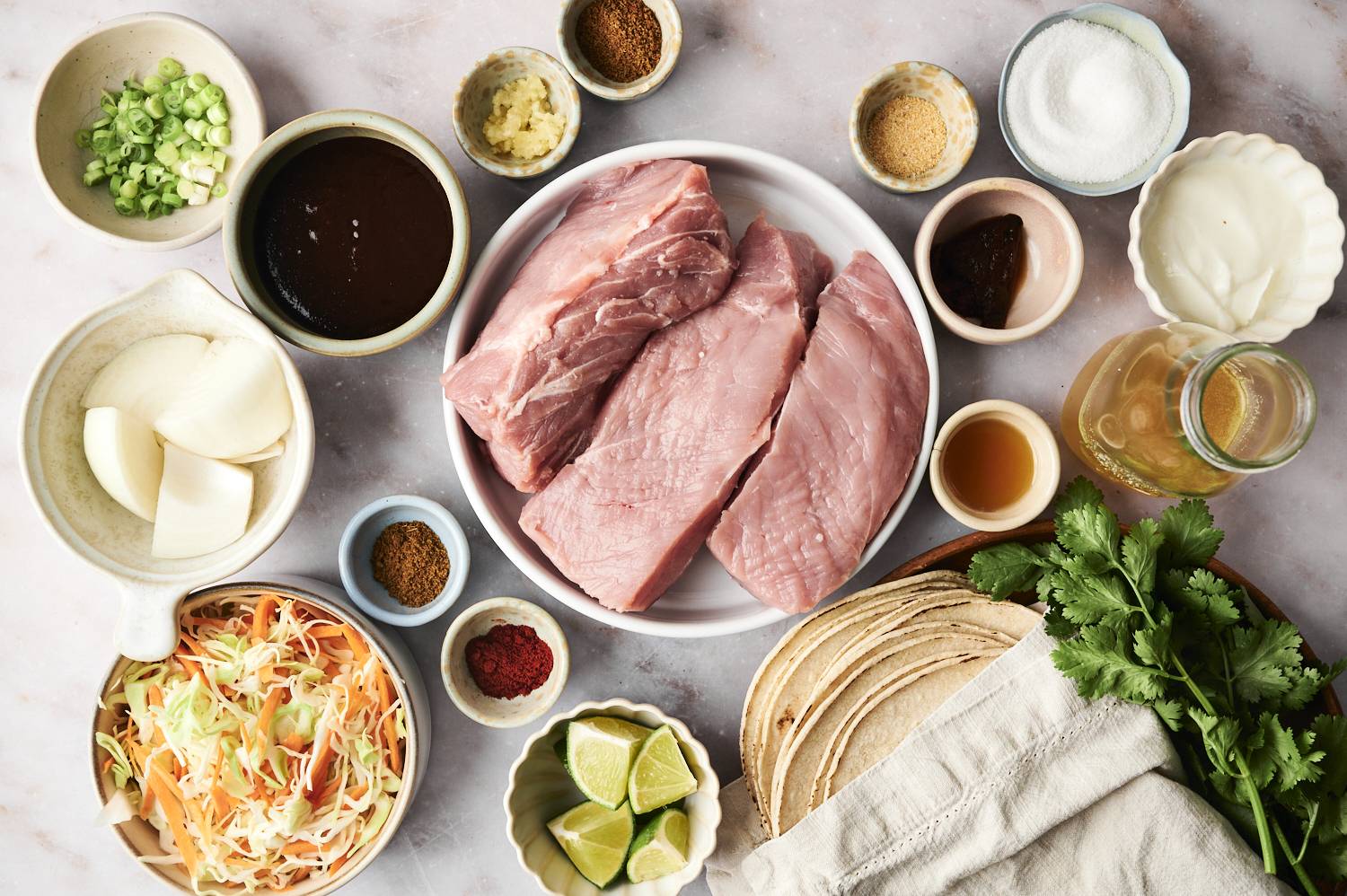 A variety of fresh ingredients arranged for a meal, including pork, tortillas, cabbage, spices, limes, and sauces on a marble surface.