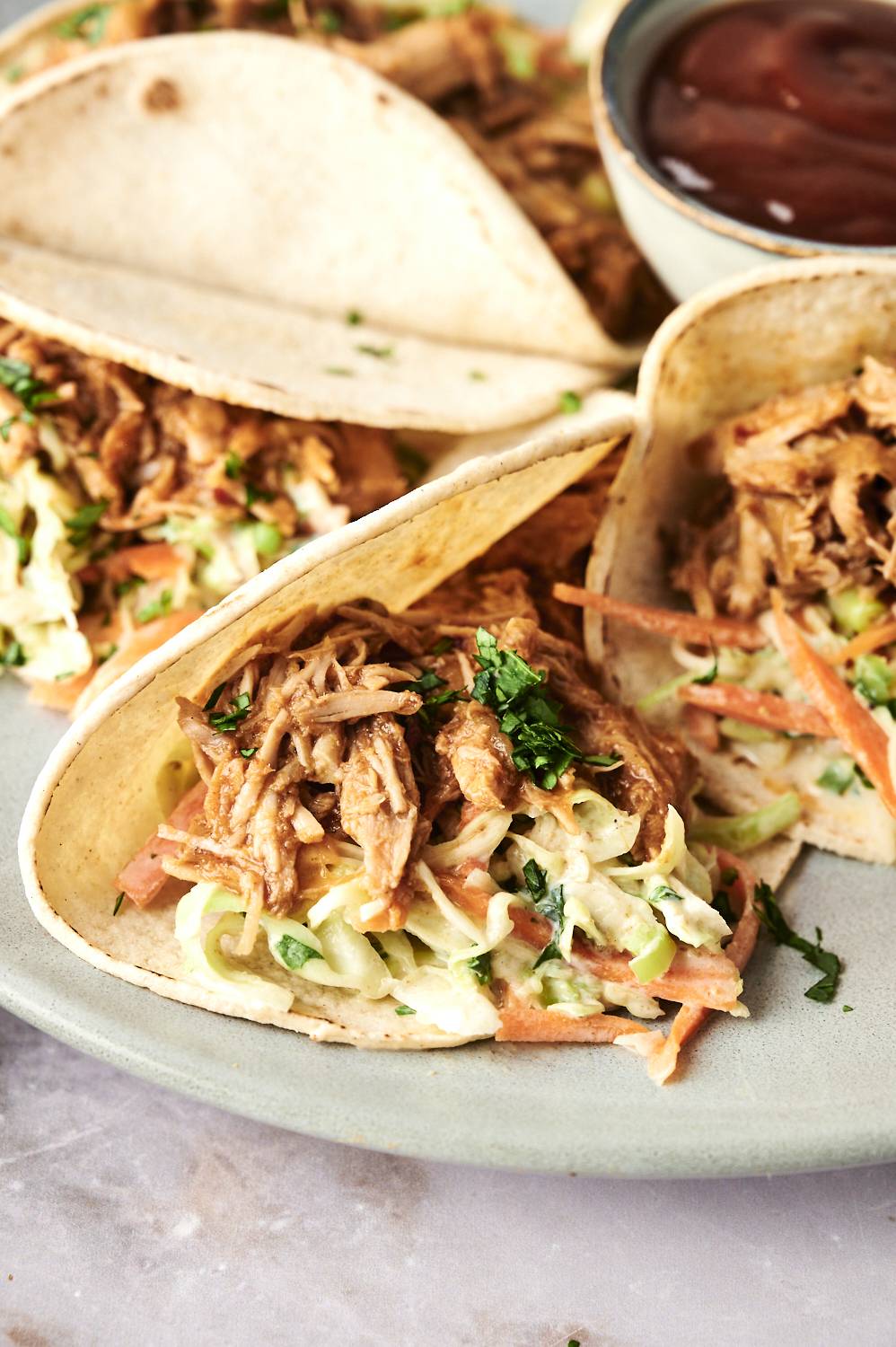 Three soft tacos filled with pulled chicken, shredded cabbage, carrots, and fresh cilantro, served on a green plate with dipping sauce.