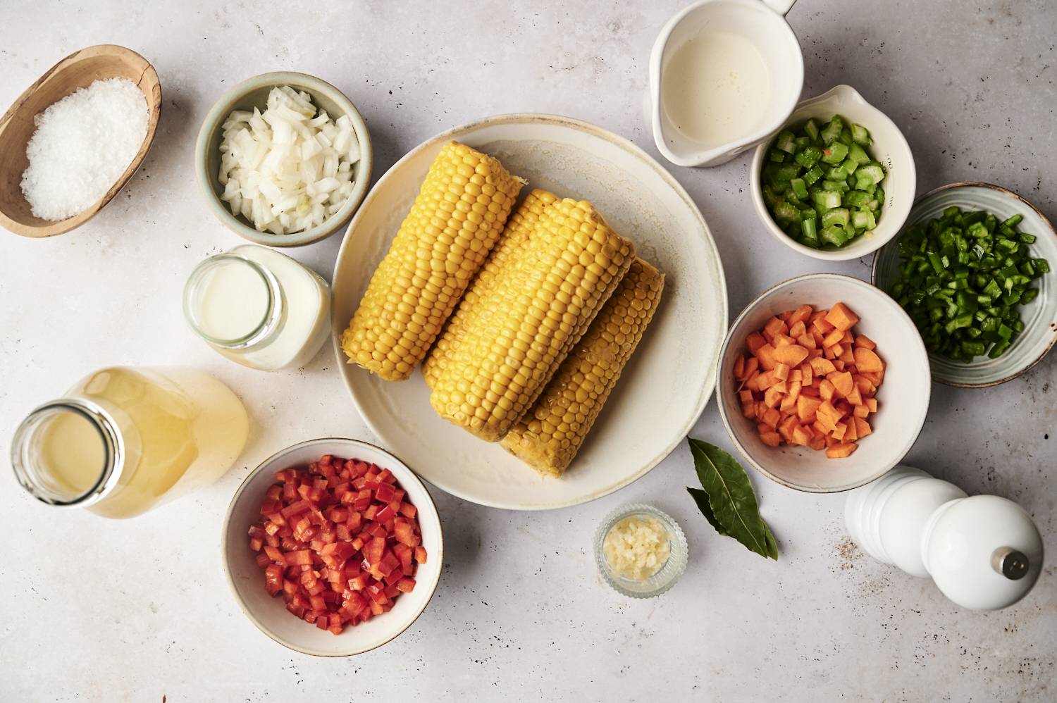 Ingredients for slow cooker roasted corn chowder, including fresh corn on the cob, diced vegetables, cream, broth, garlic, and seasonings, arranged on a light surface.