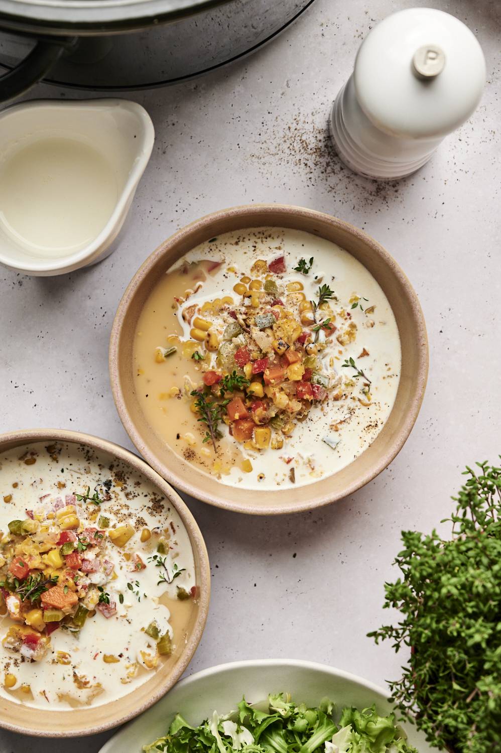 Two bowls of creamy slow cooker roasted corn chowder are garnished with roasted vegetables, fresh thyme, and black pepper, served alongside a salad.
