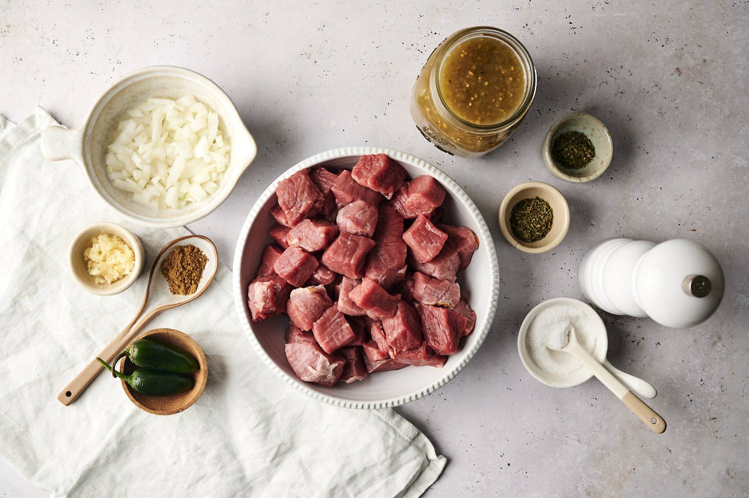 A flat-lay view of ingredients for slow cooker salsa verde beef, including cubed raw beef in a bowl, diced onions, garlic, jalapeños, spices, salt, dried herbs, and a jar of salsa verde, arranged on a neutral-toned surface.
