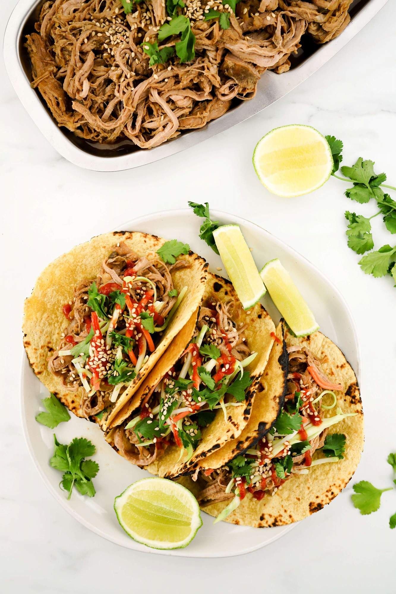 Slow Cooker Korean Pork on tortillas with slaw and Sriracha next to a plate of shredded pork.