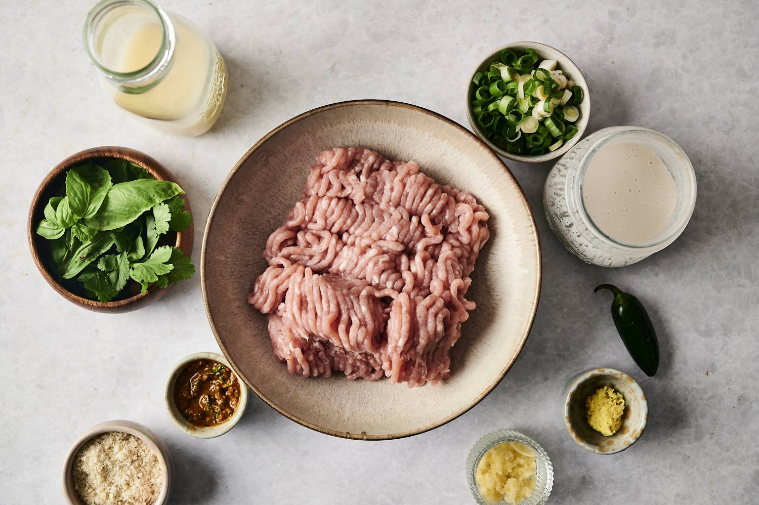 Raw ground chicken in a bowl surrounded by fresh herbs, spices, coconut milk, and aromatics.