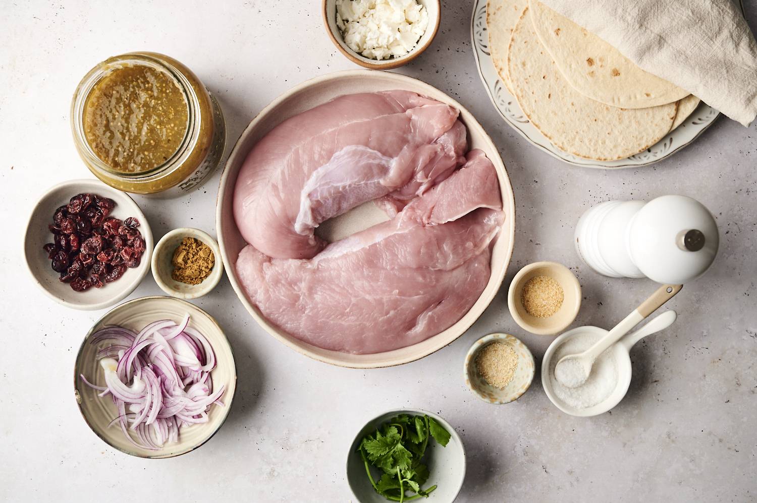 Raw turkey breast with various ingredients including tortillas, salsa, dried cranberries, red onion, spices, and fresh cilantro on a light countertop.