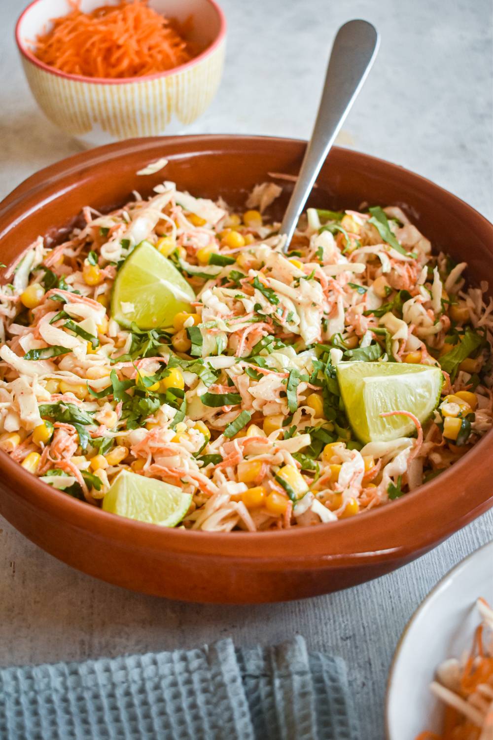 A bowl of spicy cabbage and corn slaw with lime wedges, topped with fresh cilantro. A small bowl of grated carrots sits in the background.