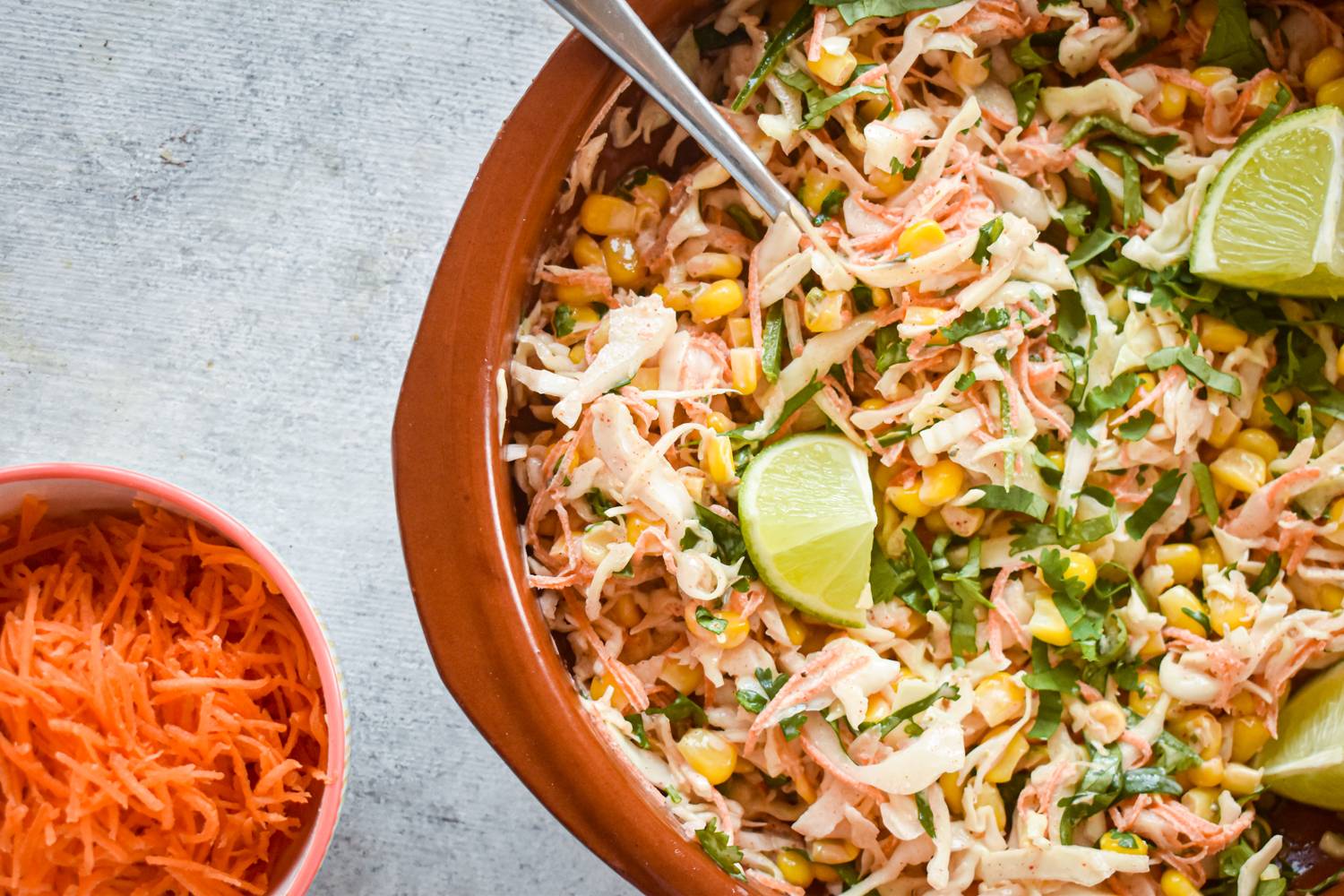 A bowl of spicy cabbage and corn slaw with lime wedges, topped with fresh cilantro. A small bowl of grated carrots sits in the background.