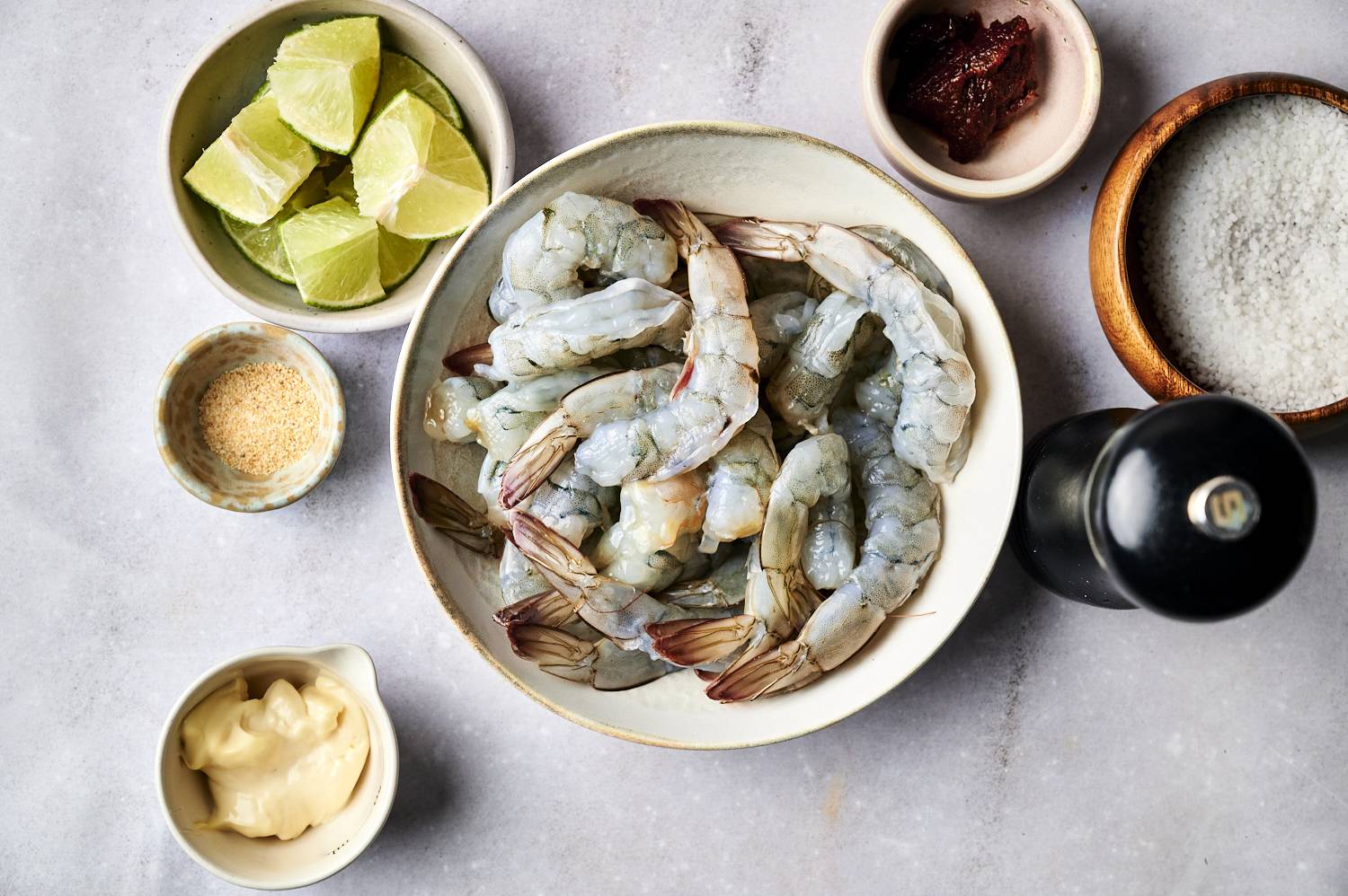 A bowl of raw shrimp surrounded by lime wedges, mayonnaise, seasonings, and salt, set against a gray countertop.