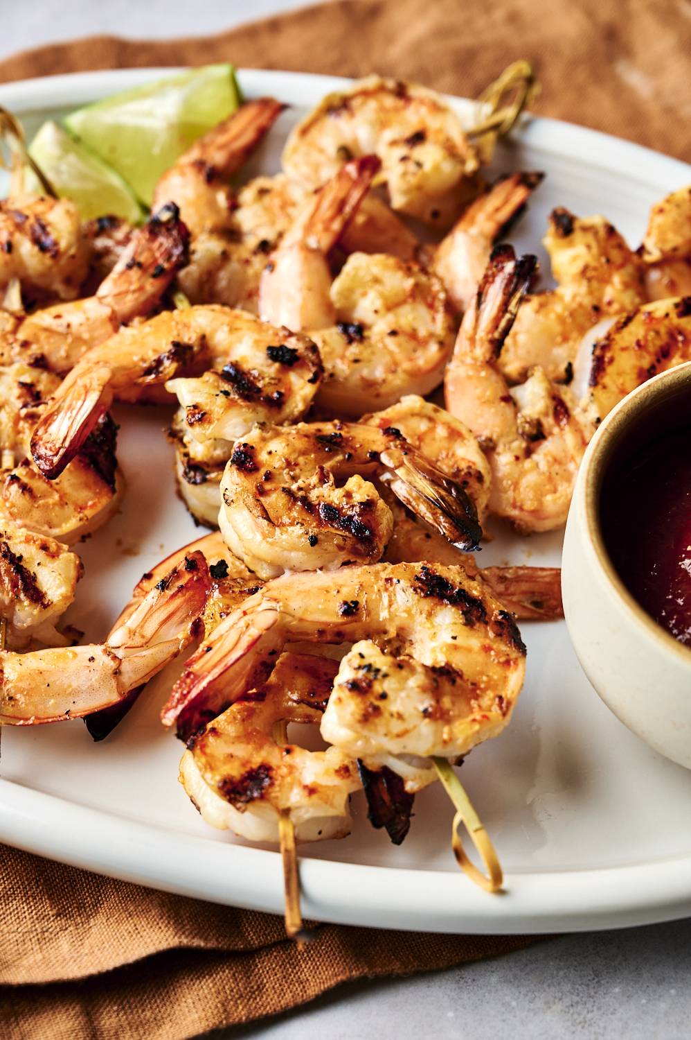 Grilled shrimp skewers glisten on a white plate, served with lime wedges and a small bowl of dipping sauce, showcasing a delicious meal.