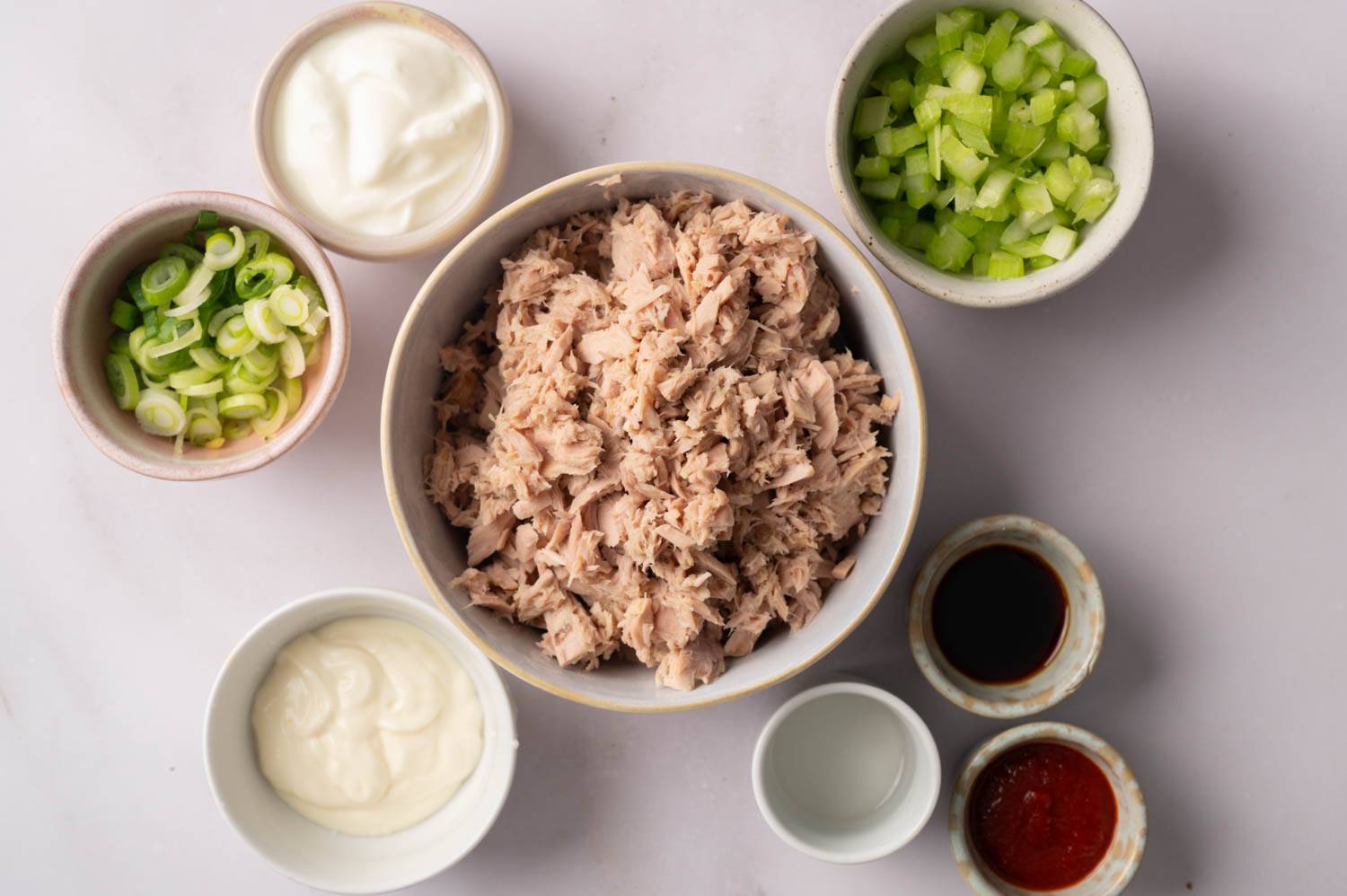 Bowls with ingredients for tuna salad on a white background. Central bowl of tuna, surrounded by green onions, celery, mayonnaise, soy sauce, and hot sauce.