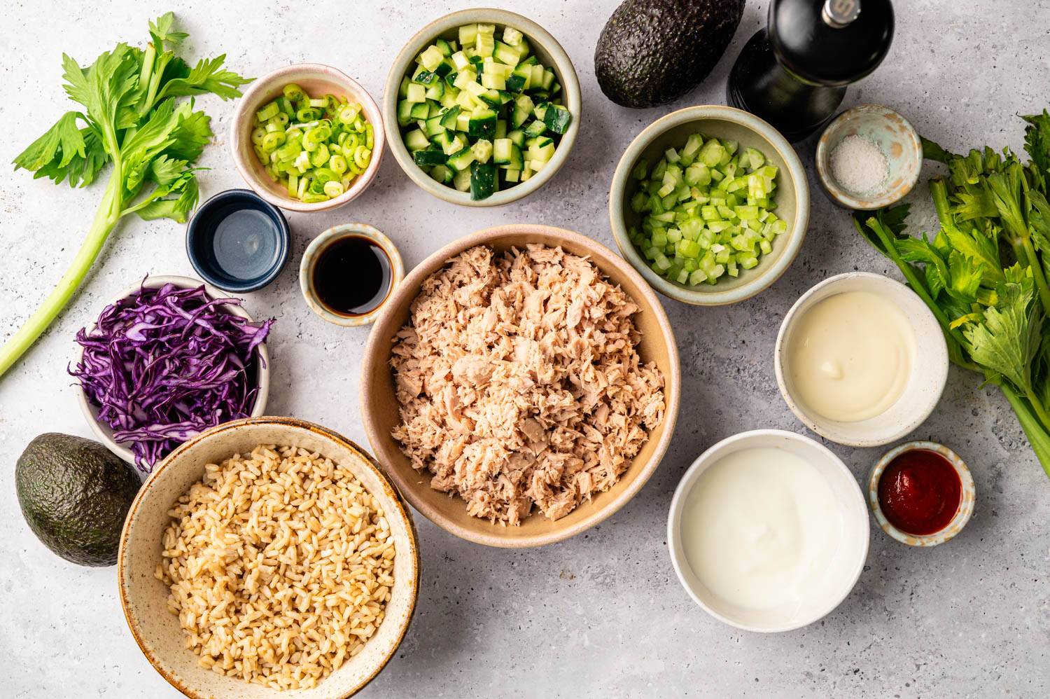 A variety of ingredients are neatly arranged, including shredded tuna, chopped cucumber, celery, green onions, purple cabbage, avocado, rice, soy sauce, and sauces, ready for meal prep. The tone is fresh and vibrant.