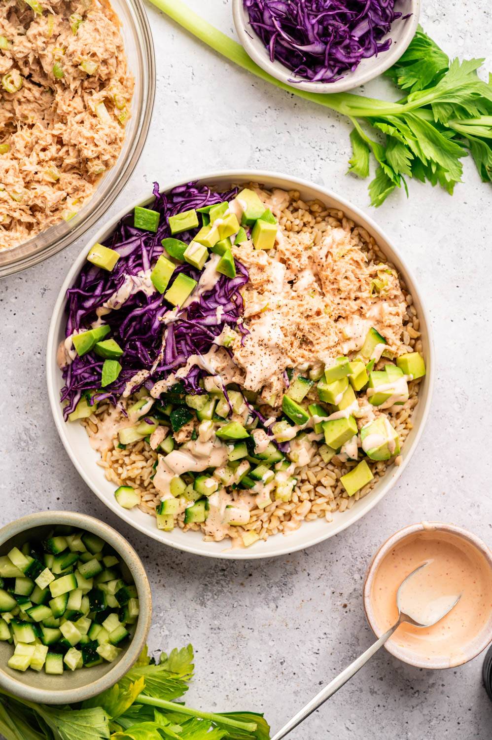 A colorful rice bowl with tuna, diced avocado, purple cabbage, and sauce on a gray surface. Nearby, bowls of tuna, cucumbers, and dressing are visible.