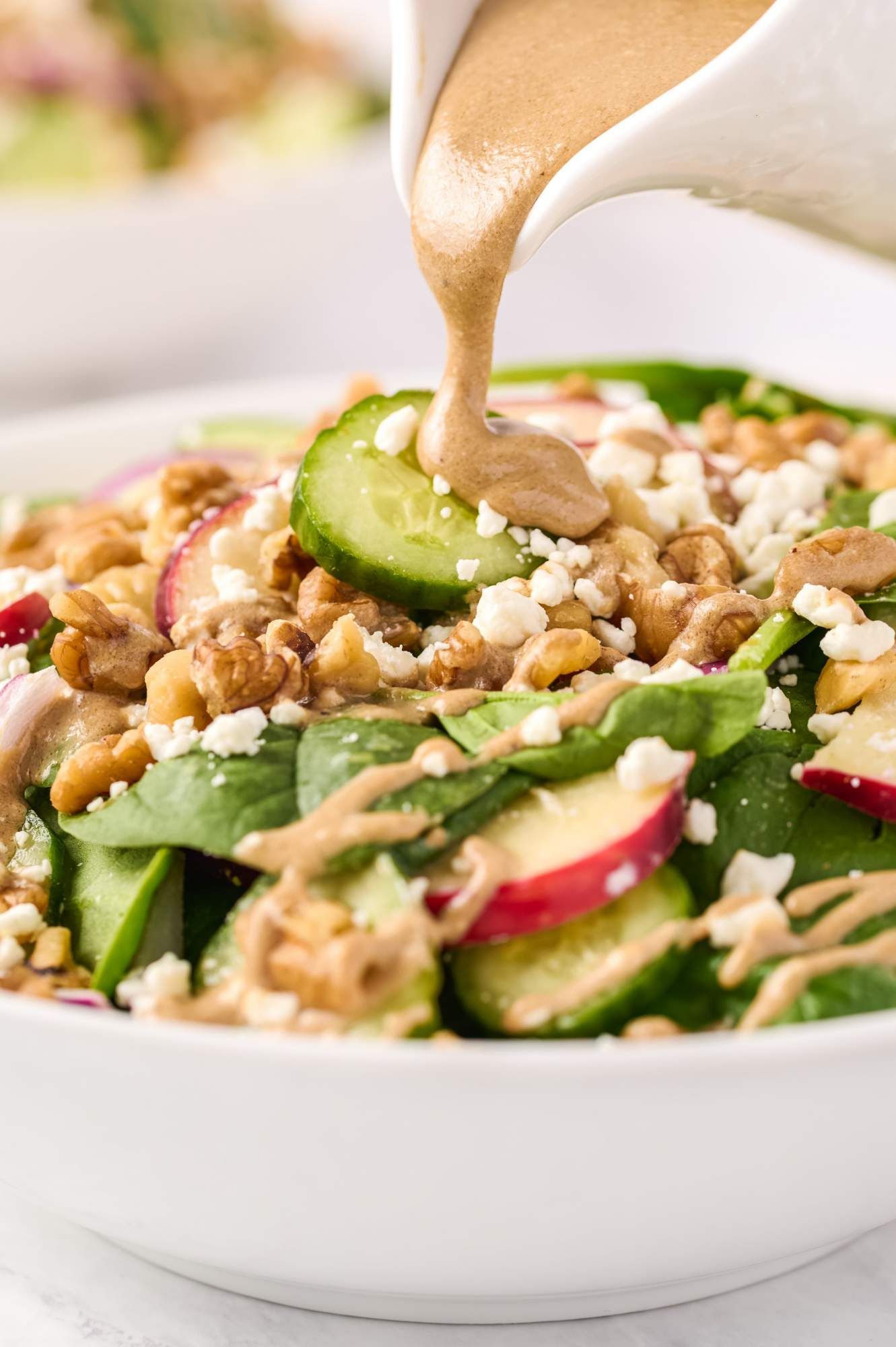 Balsamic vinaigrette being poured over spinach salad with apples, goat cheese, and walnuts.