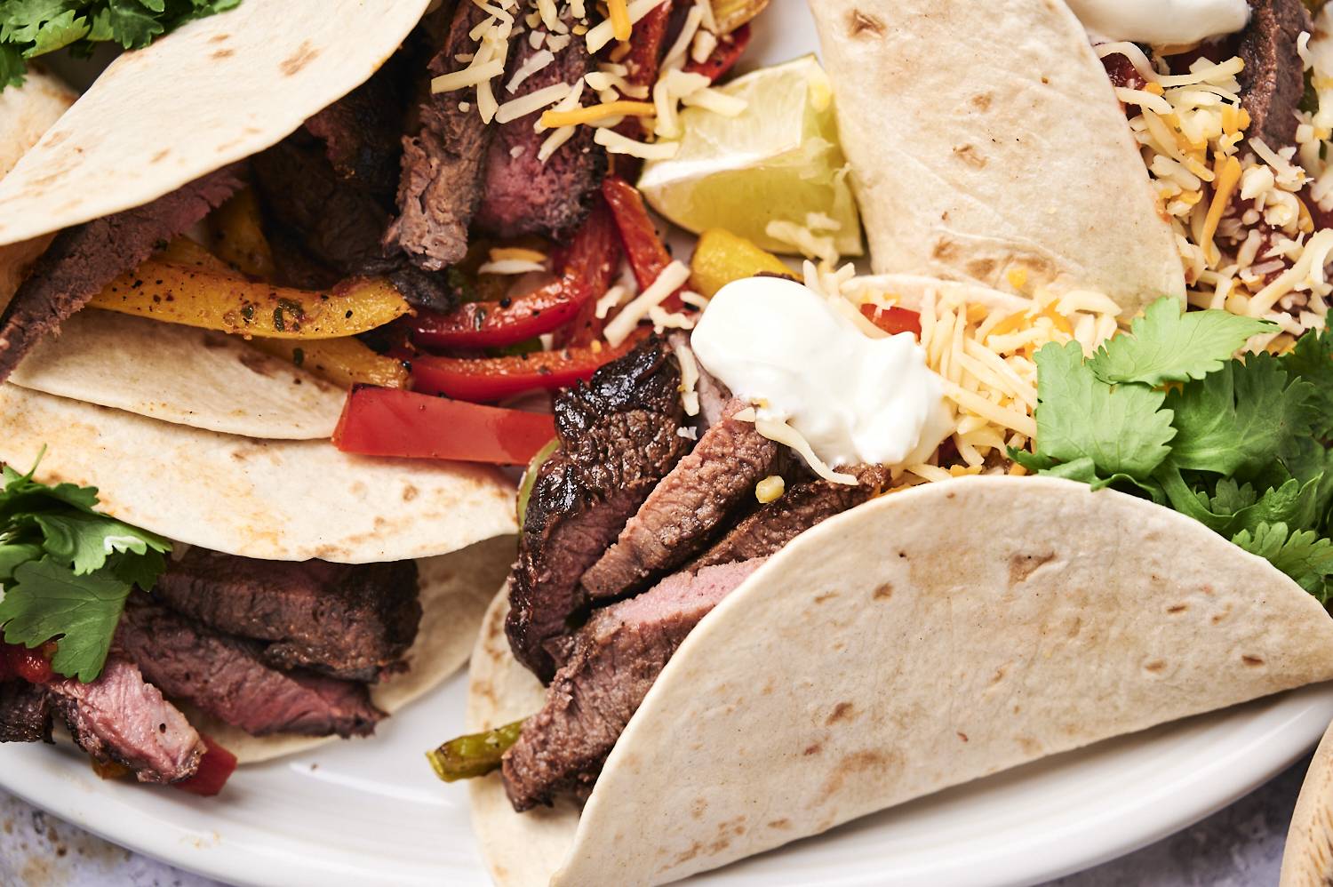 Beef fajitas served in flour tortillas with shredded cheese, cilantro, sour cream, and lime wedges.