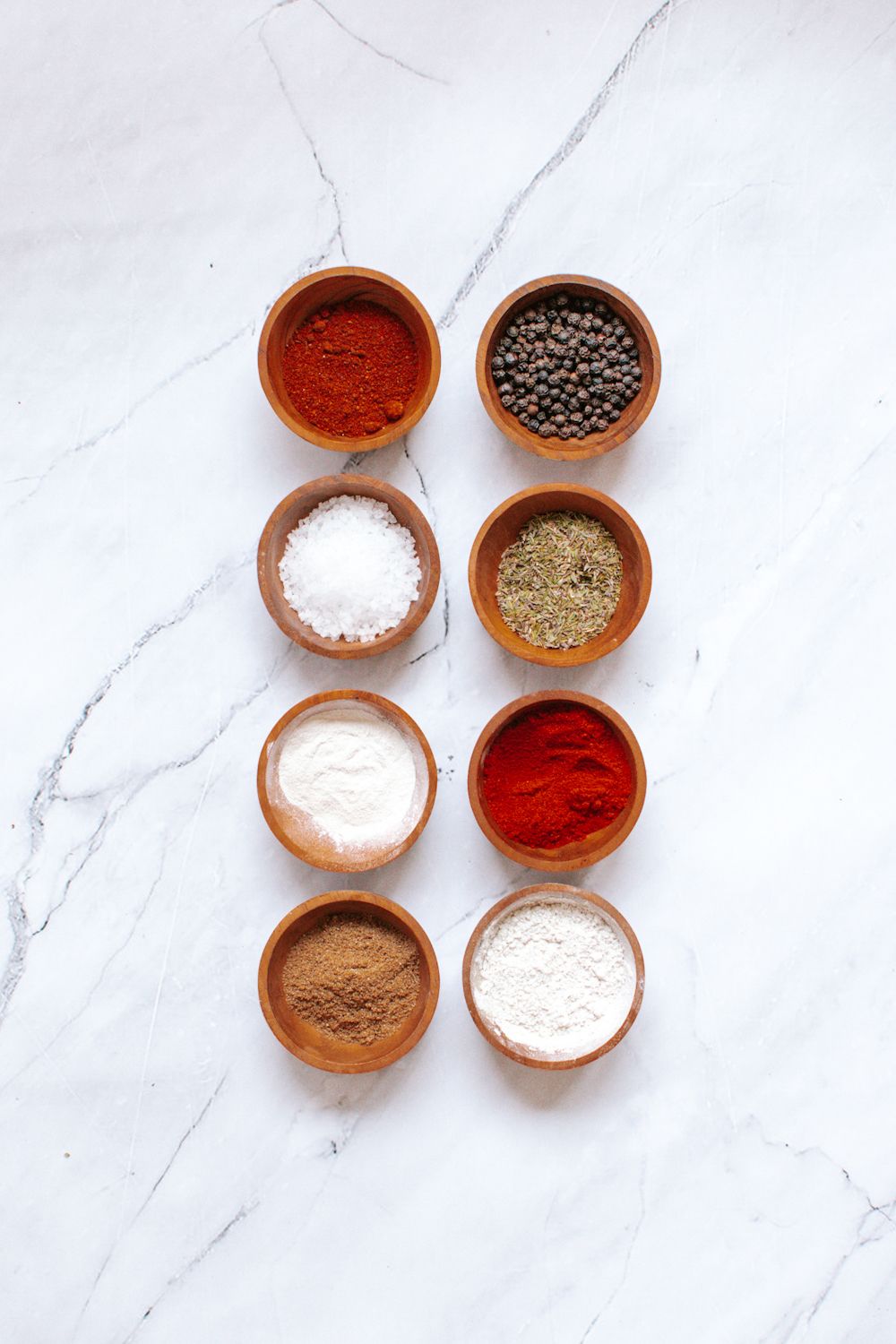 Ingredients for steak seasoning in small wooden bowls including paprika, peppercorns, kosher salt, fennel, onion powder, garlic powder, cumin, and chili powder.