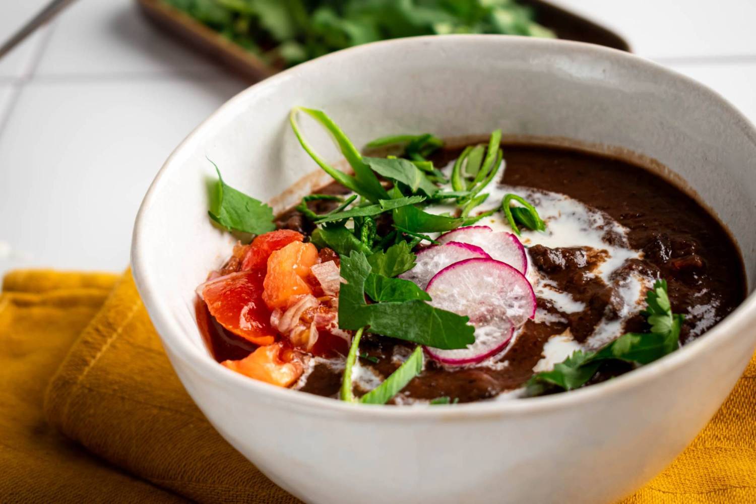 A bowl of black bean soup topped with sliced radishes, fresh herbs, and a drizzle of cream. The rich soup is garnished with vibrant vegetables.