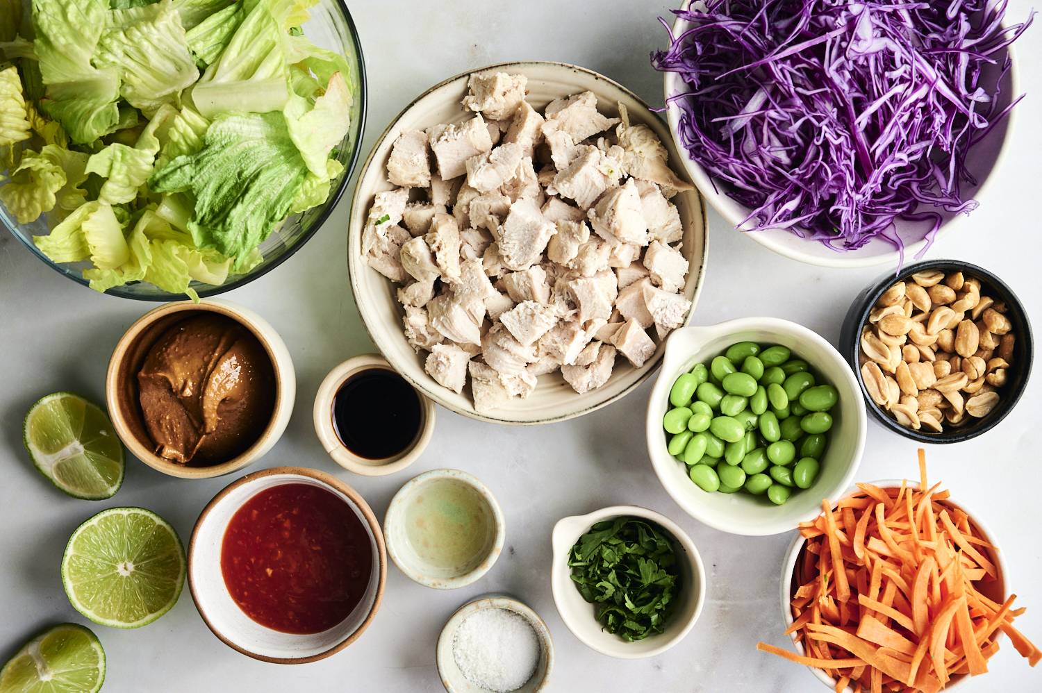 A colorful arrangement of salad ingredients: romaine lettuce, shredded purple cabbage, diced chicken, carrots, edamame, peanuts, and sauces.
