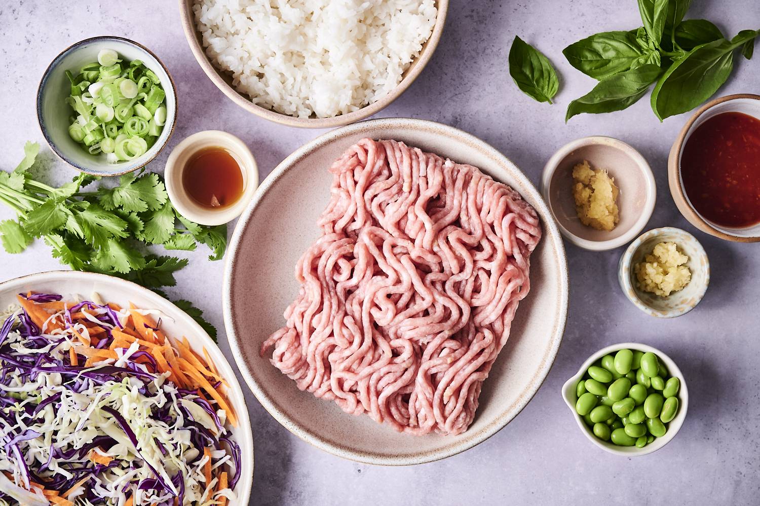 An overhead view of ingredients for a meal, featuring ground meat, rice, fresh herbs, chopped vegetables, sauces, and edamame in bowls.