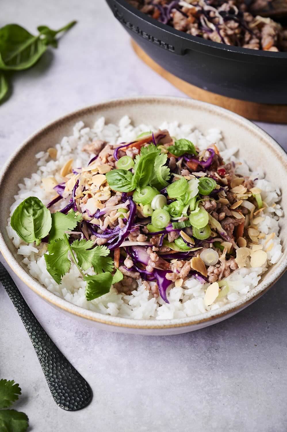 Bowl of white rice topped with sautéed ground meat, red cabbage, scallions, and fresh herbs, served beside a dark pot.