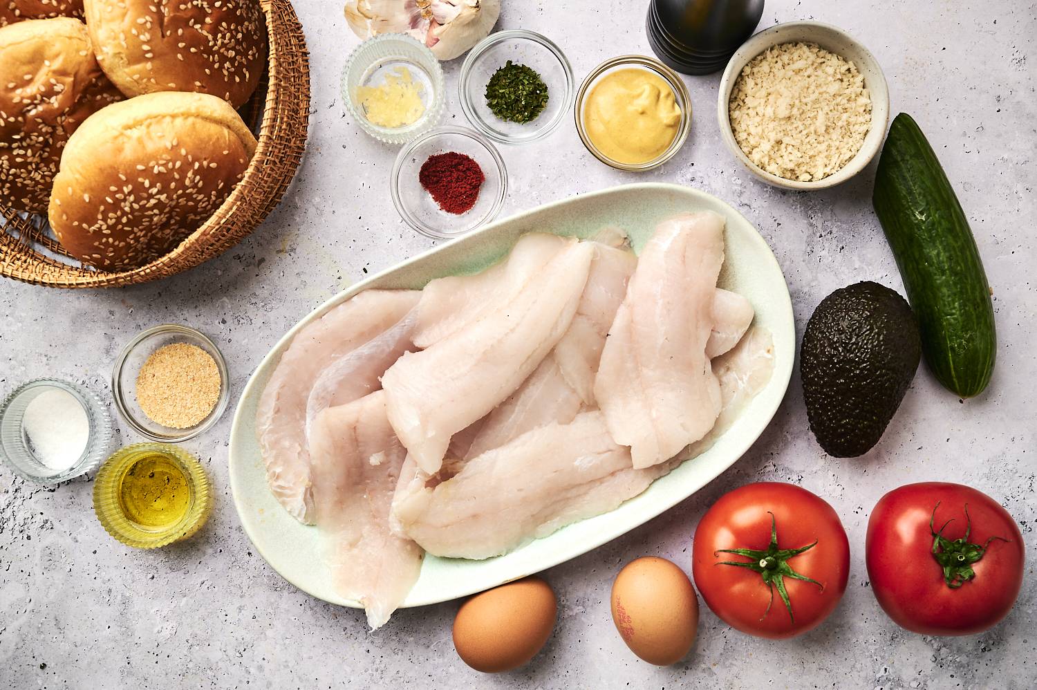 Ingredients for tilapia fish burgers include raw tilapia fillets, eggs, tomatoes, cucumber, avocado, bread crumbs, spices, mustard, garlic, and sesame seed buns.