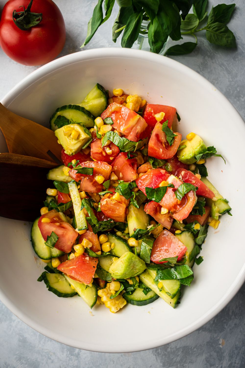 Cucumber tomato salad with chopped tomatoes, corn, avocado, corn, and basil in a bowl with wooden tongs.