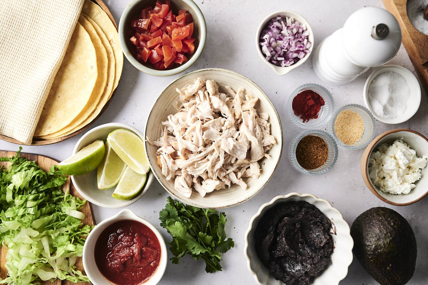 Ingredients for making tostadas, including tortillas, shredded chicken, diced tomatoes, onions, black beans, avocado, lime wedges, lettuce, cilantro, and spices such as chili powder, garlic powder, and salt. There is also crumbled cheese and salsa on the table.