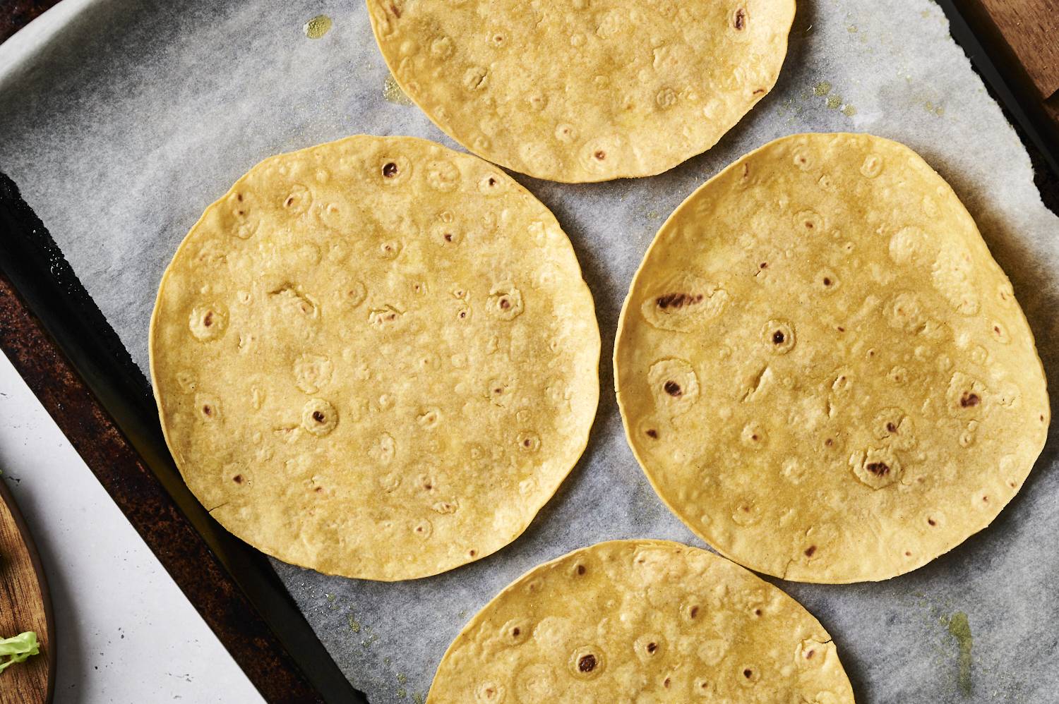 Four plain tortillas arranged on a baking sheet lined with parchment paper. They are ready to be toasted or baked for making tostadas.