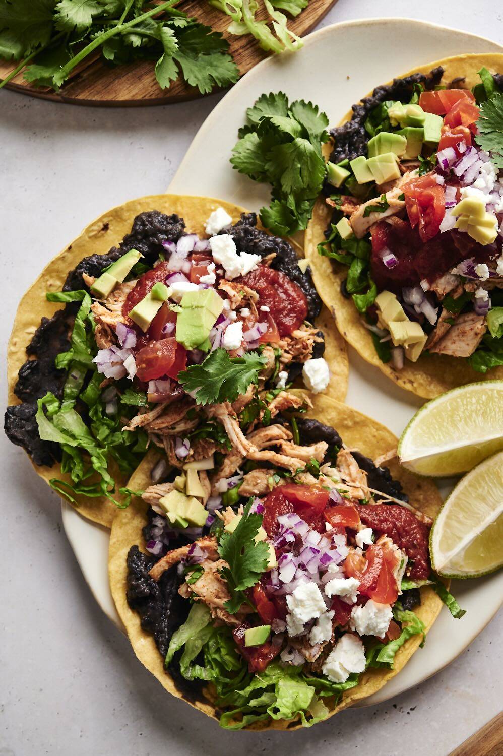 Tostadas topped with shredded chicken, black beans, fresh lettuce, avocado, diced tomatoes, red onions, cilantro, and crumbled cheese, with a wedge of lime on the side.