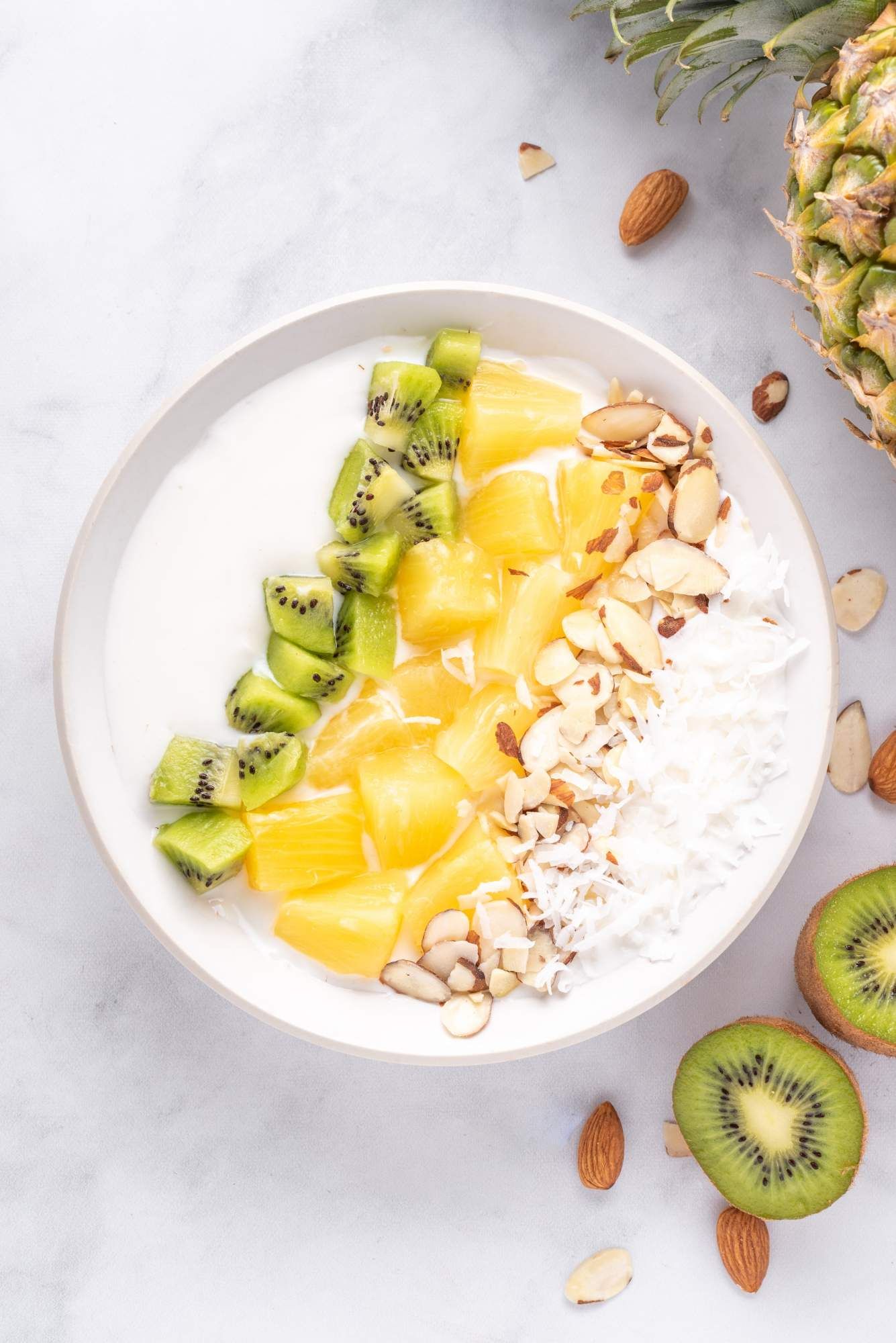 Yogurt bowl with pineapple, kiwi, shredded coconut, and sliced almond in a white bowl with kiwi on the side.