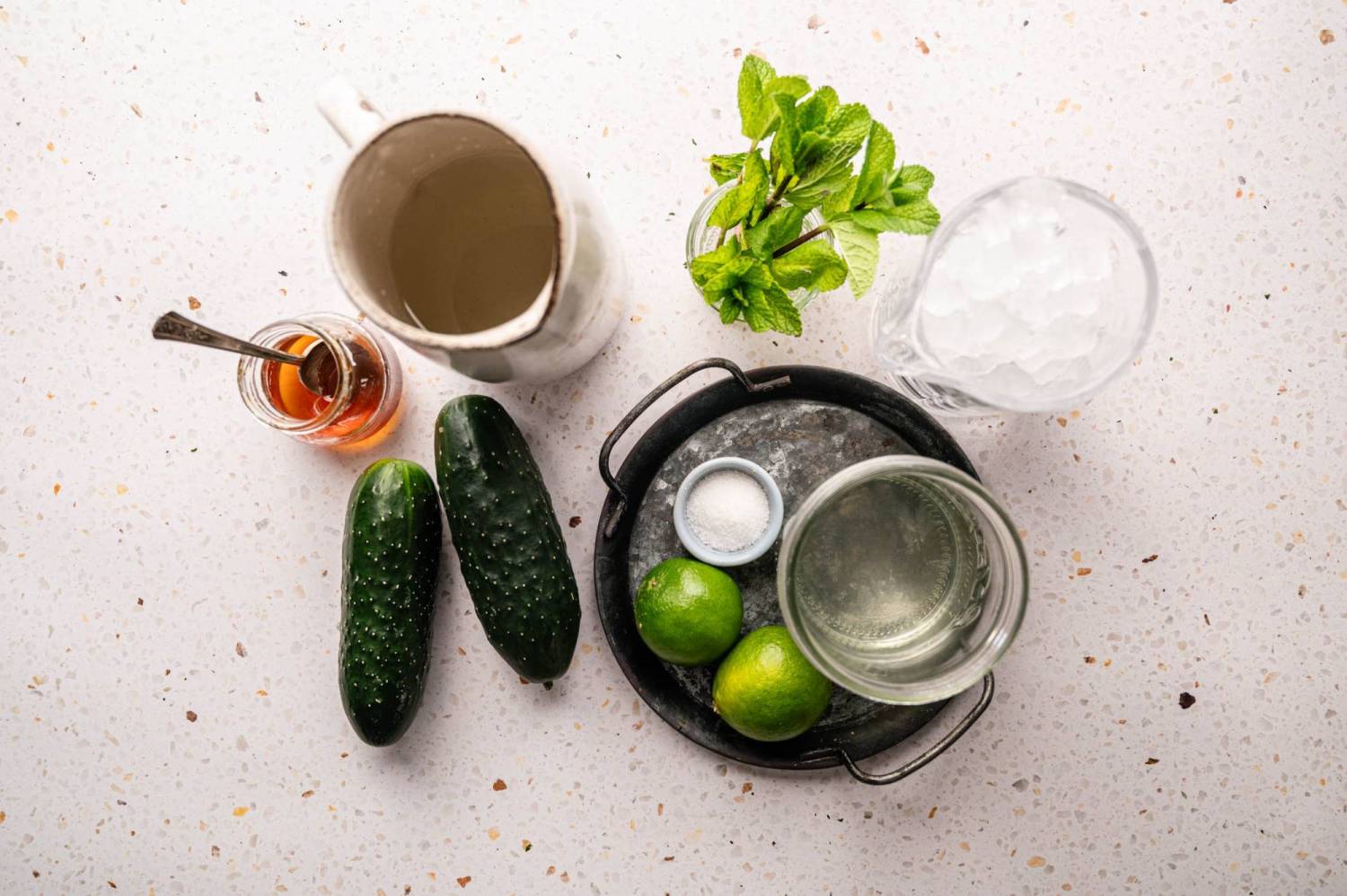 Ingredients for a refreshing drink are arranged on a speckled surface: cucumbers, honey, mint, ice, limes, water, and salt in a small dish.