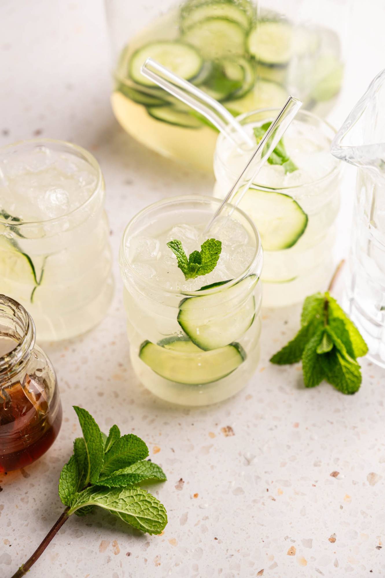 Refreshing drinks with cucumber slices, mint sprigs, and ice in clear glasses on a light speckled surface, conveying a fresh, summery feel.