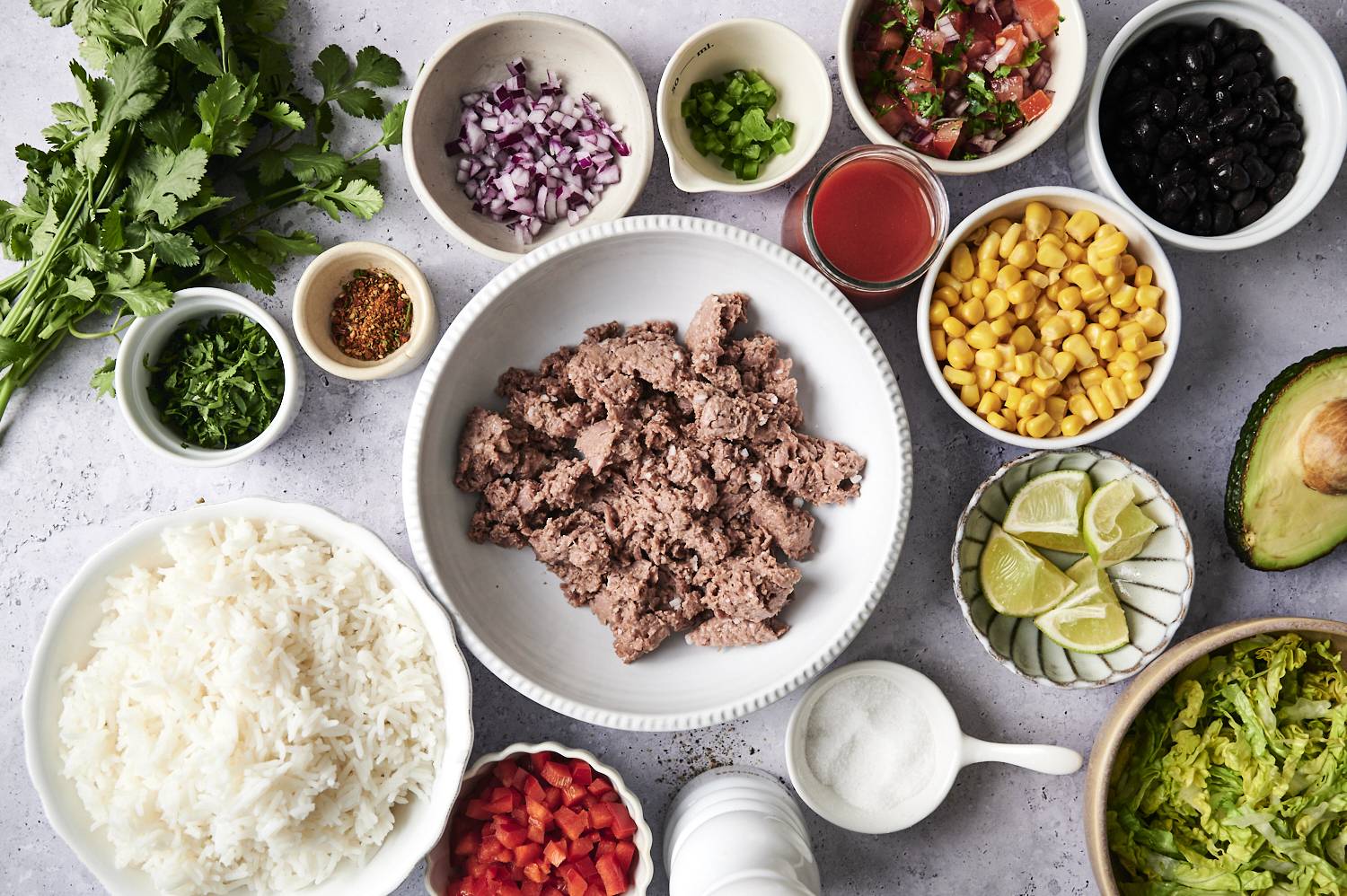 Ingredients for vegan ground beef burrito bowls, including plant-based ground beef, rice, black beans, corn, diced vegetables, avocado, lime wedges, fresh herbs, and seasonings, arranged on a light surface.