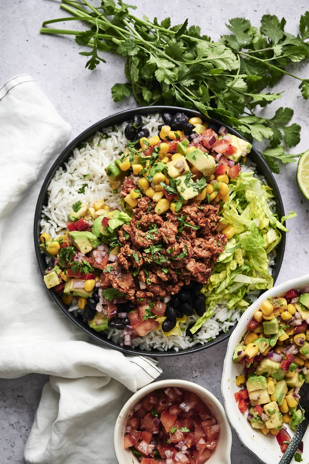 A vegan ground beef burrito bowl is served with cilantro lime rice, black beans, seasoned plant-based ground beef, lettuce, corn, avocado, and fresh pico de gallo, garnished with chopped cilantro.