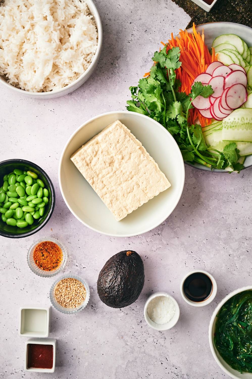 Ingredients for vegetarian sushi bowls including rice, edamame, avocado, carrots, cucumbers, seaweed, tofu, and spicy mayo.