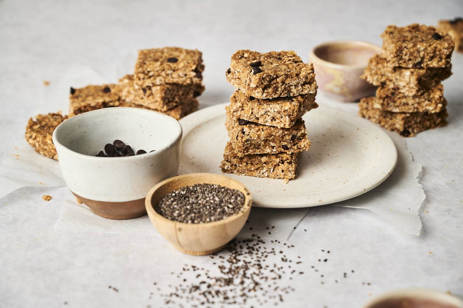 A plate piled with homemade oat bars, surrounded by bowls of chocolate chips and chia seeds on a light background.
